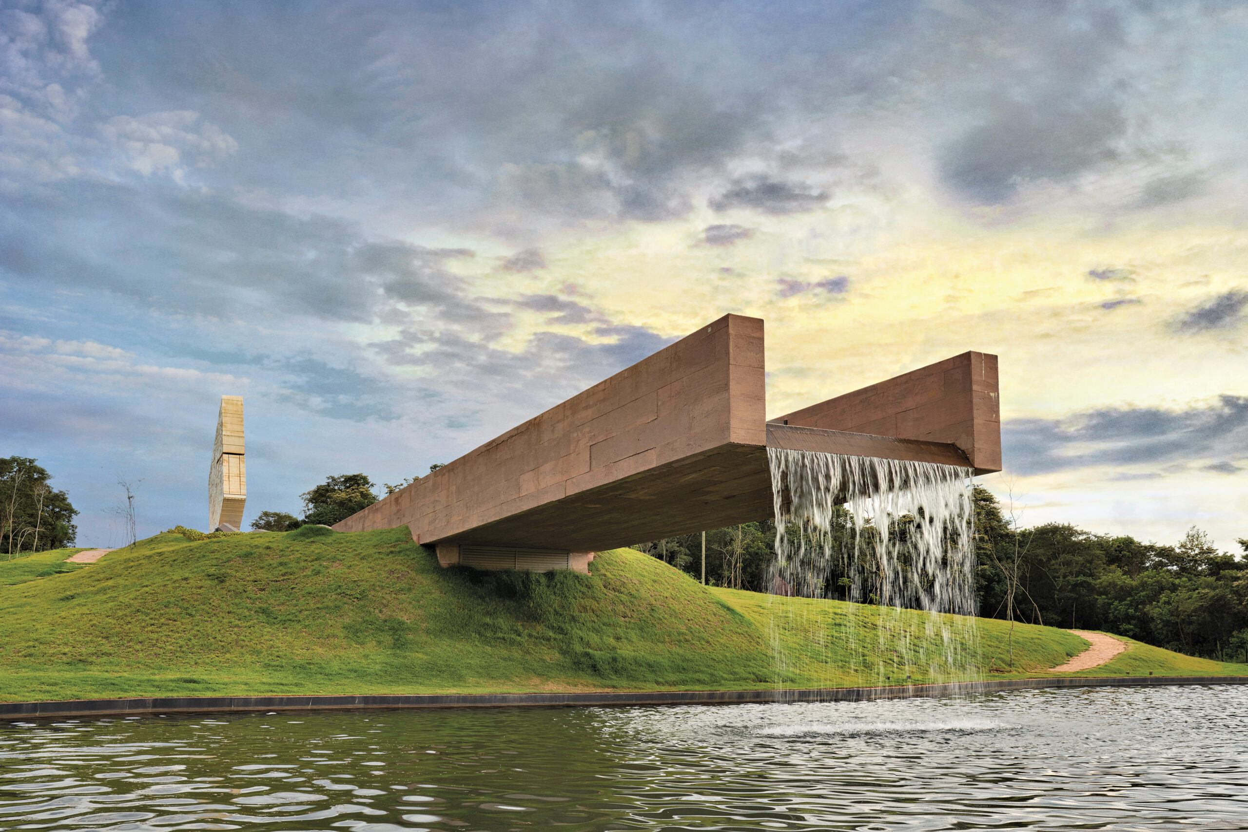 On a grassy knoll in front of a waterway, the Brumadinho Memorial extends a cantilevered arm over the water, flowing with rushing water.