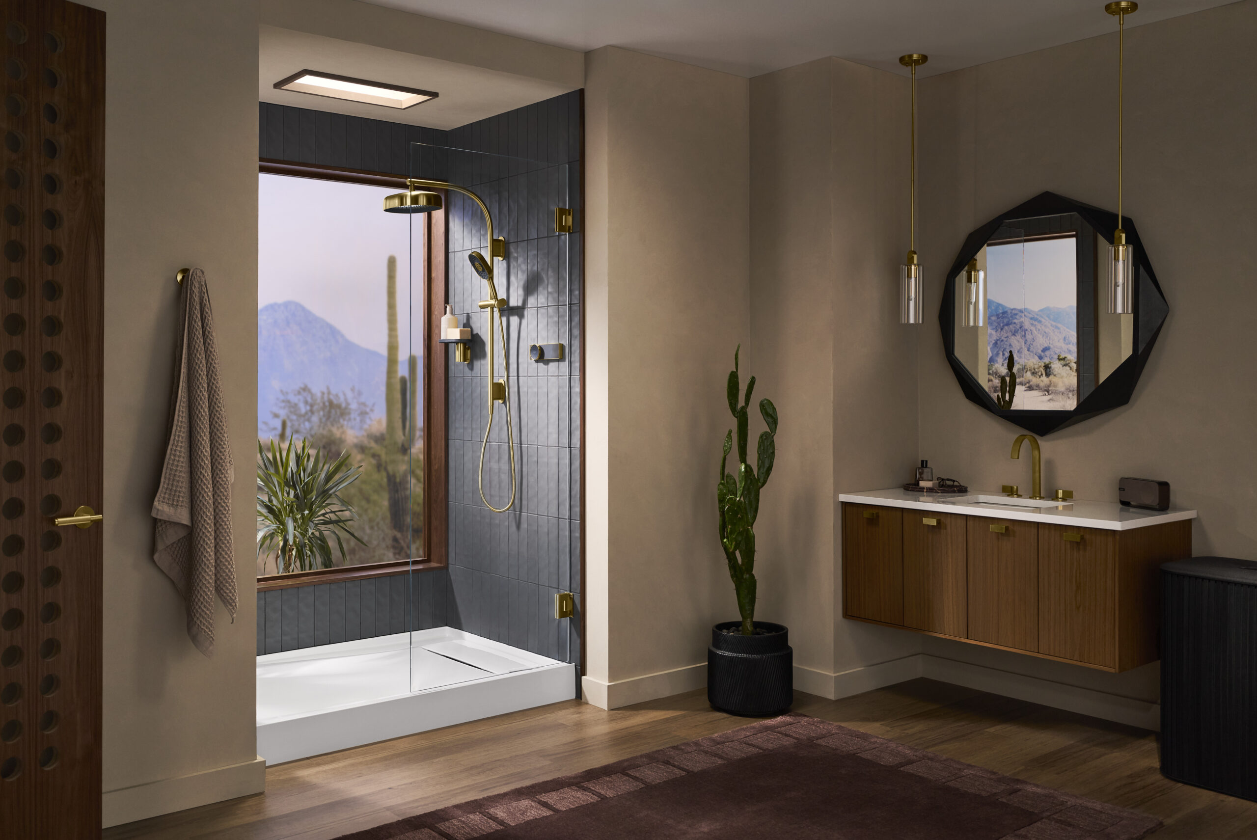 Modern bathroom with a glass shower, large window showing desert landscape, cactus plant, floating wood vanity with mirror, and pendant lights.