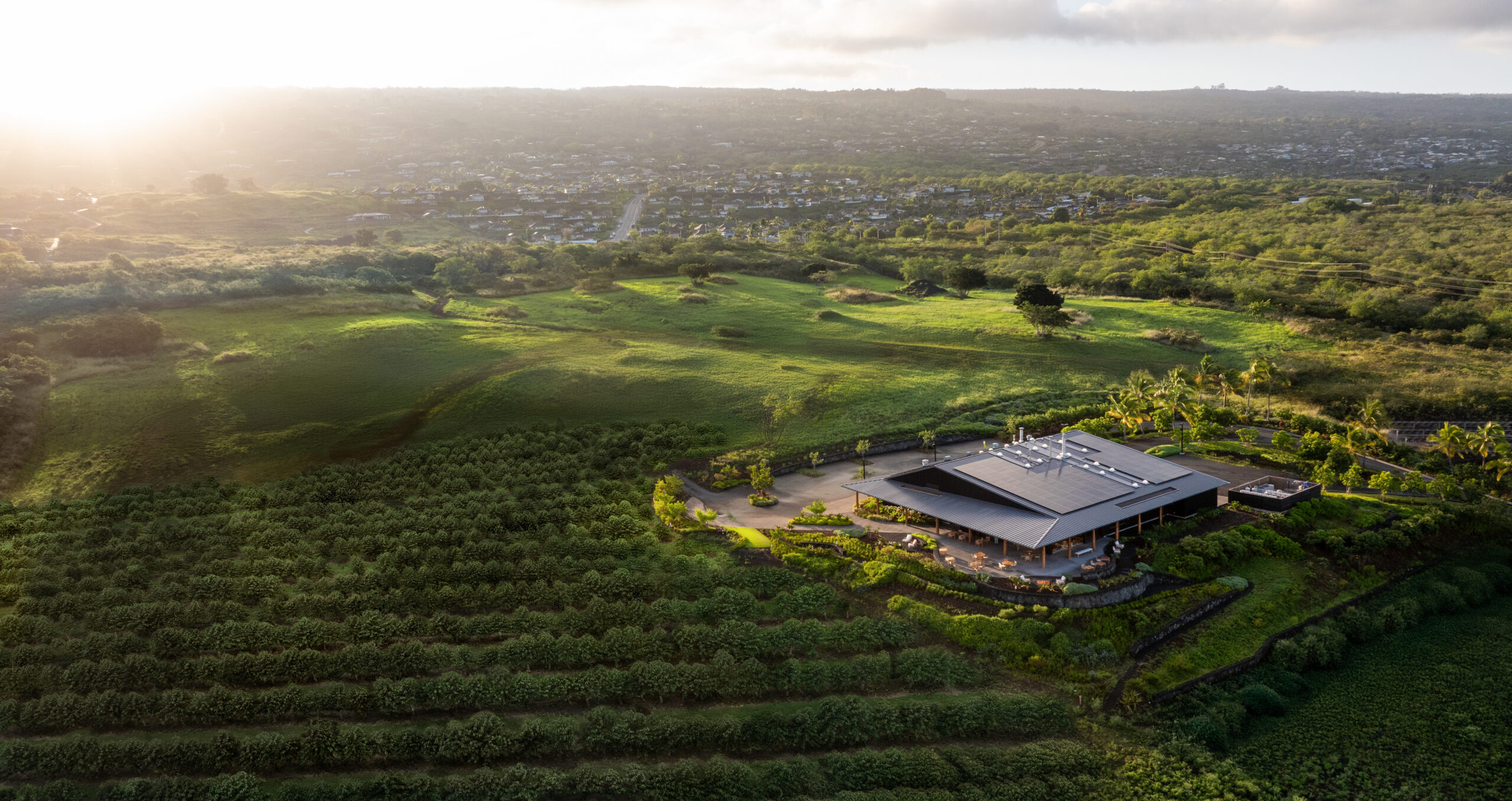 Aerial view of a modern building surrounded by green fields and hills, with a city in the distance under a cloudy sky at sunrise or sunset.
