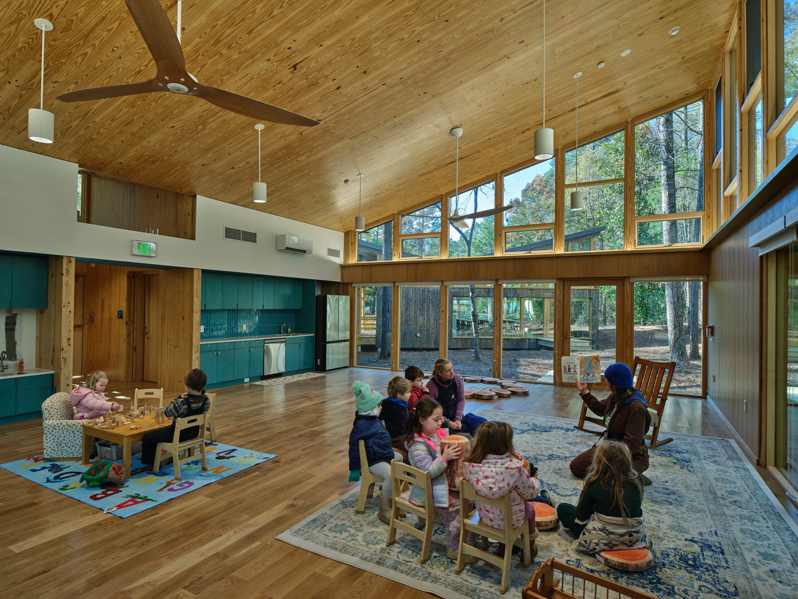 Children sit in groups playing and listening to an adult read in a spacious, wood-paneled room with large windows and natural light.