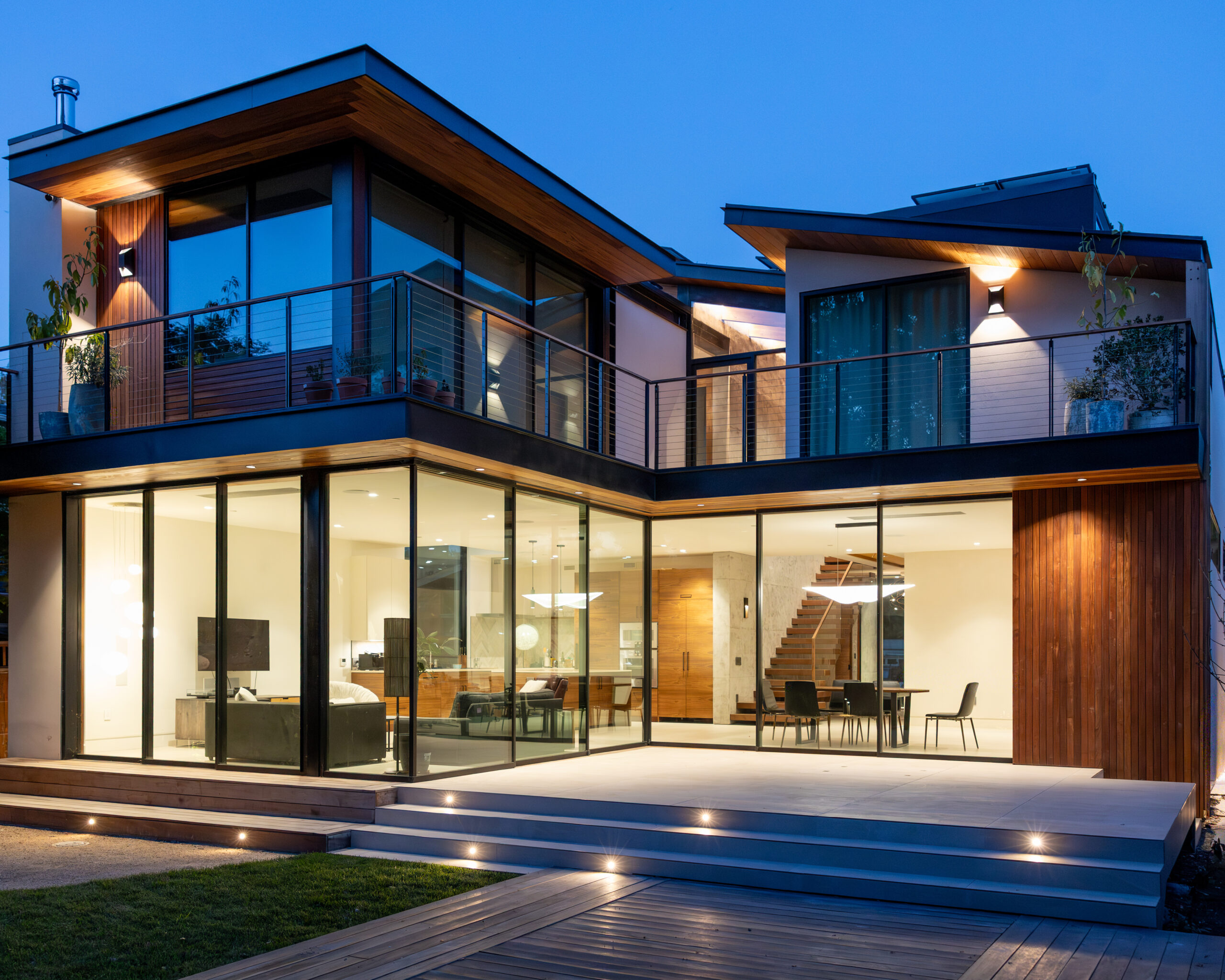 Modern two-story house with large glass windows, wooden accents, and an outdoor patio illuminated at dusk. The interior shows a living area, dining table, and staircase.
