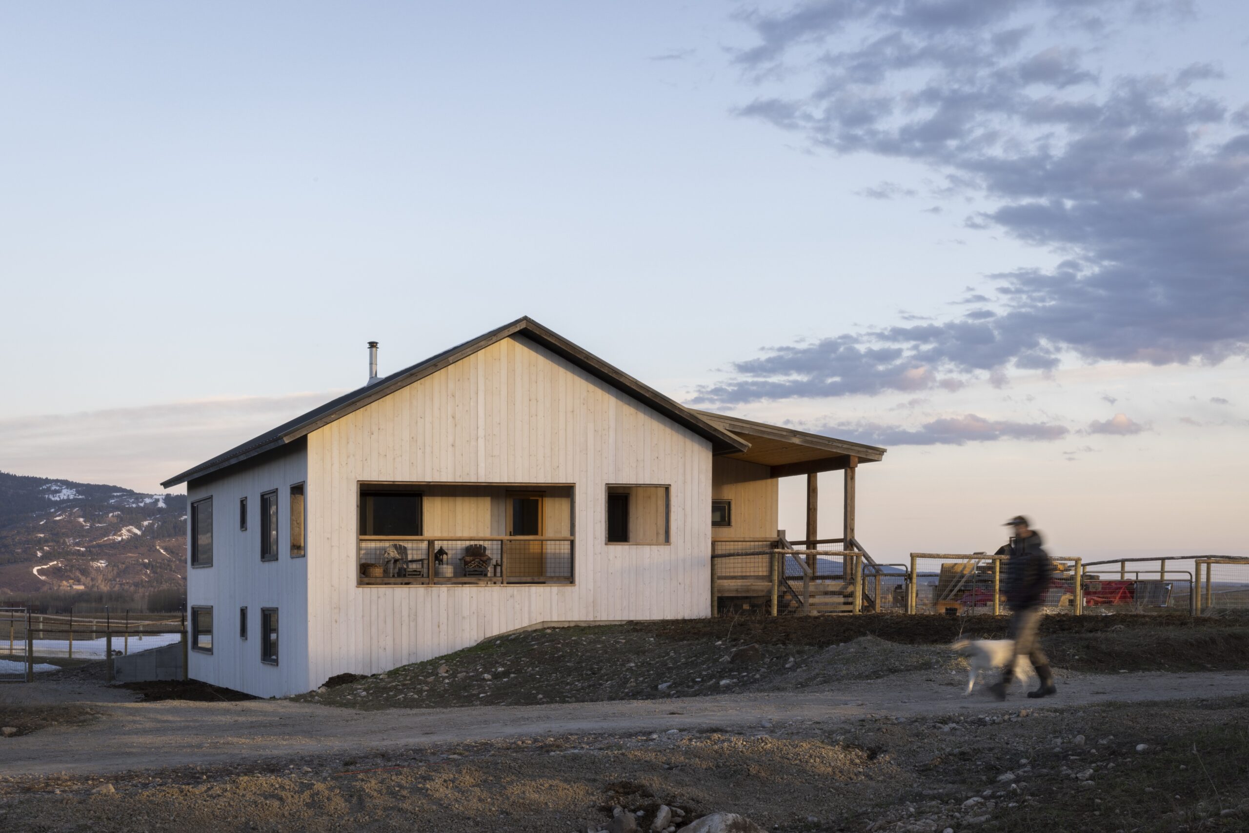 A modern white house with a covered porch sits on a dirt road at sunset; a person walks a dog in the foreground, with mountains and a partly cloudy sky in the background.