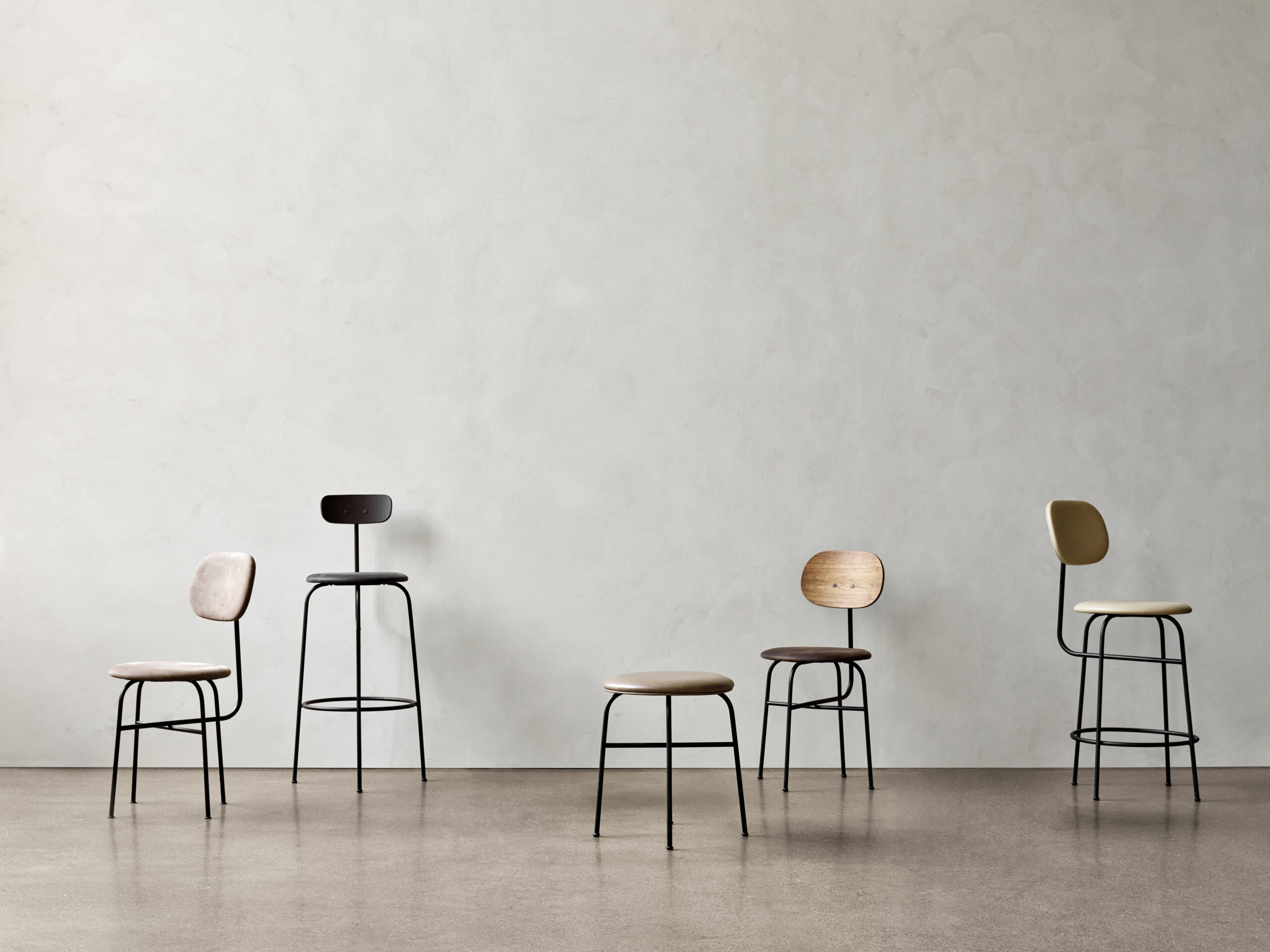 Six modern chairs and stools with minimalist silhouettes stand in front of a grey, lime-washed wall. 