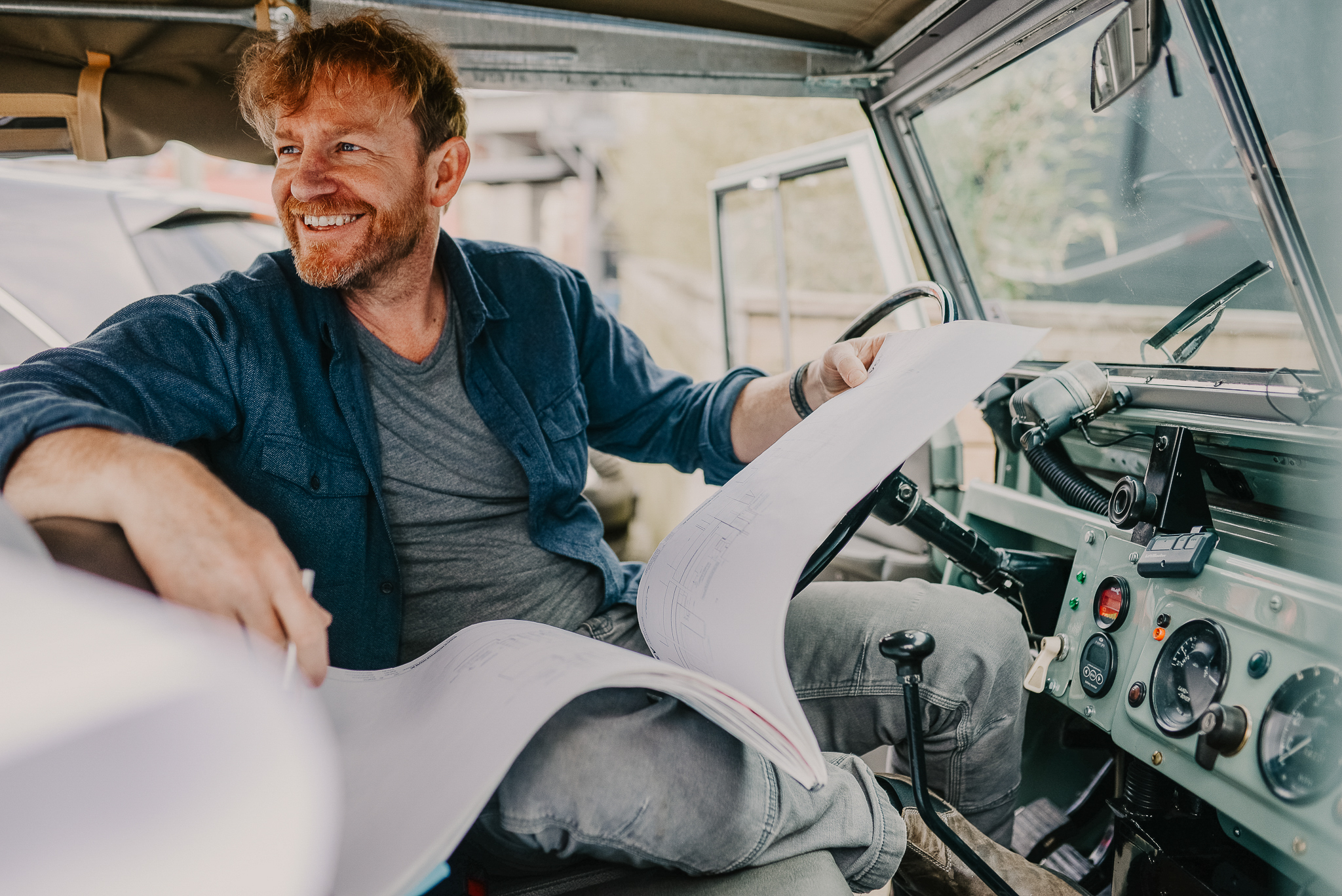 Smiling man sits in a vehicle, holding large architectural blueprints across his lap and looking out the window—perhaps reflecting on innovative designs inspired by Announcing the Winners of the 2026 METROPOLIS Planet Positive Awards.