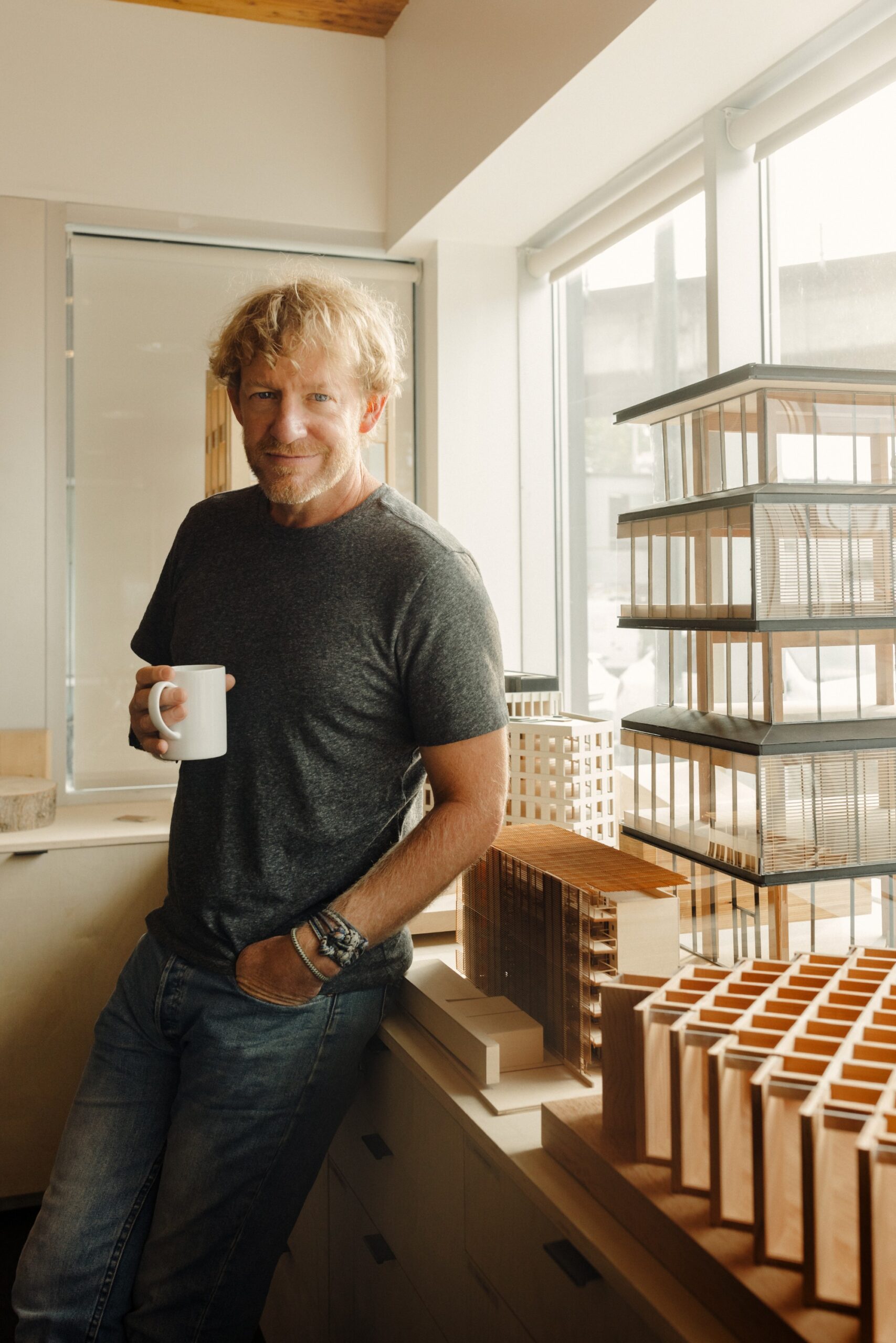 A man, Michael Green, in a gray shirt and jeans holds a white mug and stands by a window next to several architectural models made of wood and clear materials.