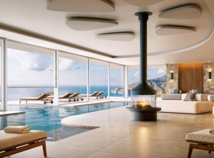 19 Products to Specify According to Design Pros: Modern indoor pool lounge with a central fireplace, white sofas, and floor-to-ceiling windows overlooking a coastal view with mountains and ocean.