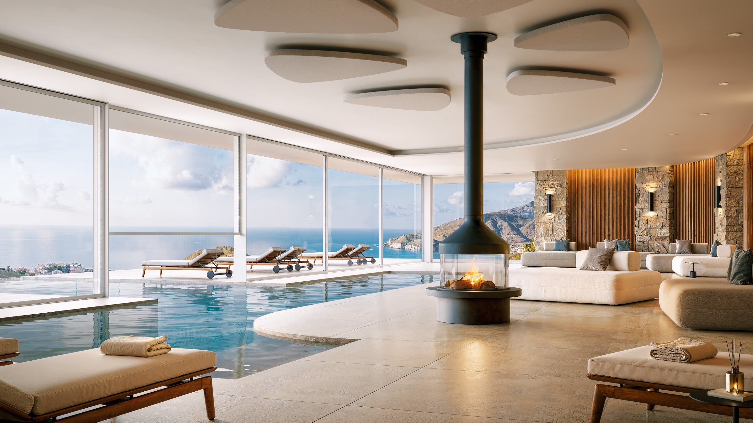 Products to Specify: Modern indoor pool lounge with a central fireplace, white sofas, and floor-to-ceiling windows overlooking a coastal view with mountains and ocean.