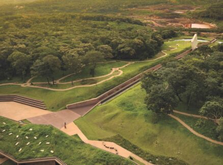 Aerial view of a modern building with geometric lines, surrounded by winding paths and dense green trees in a park-like landscape.