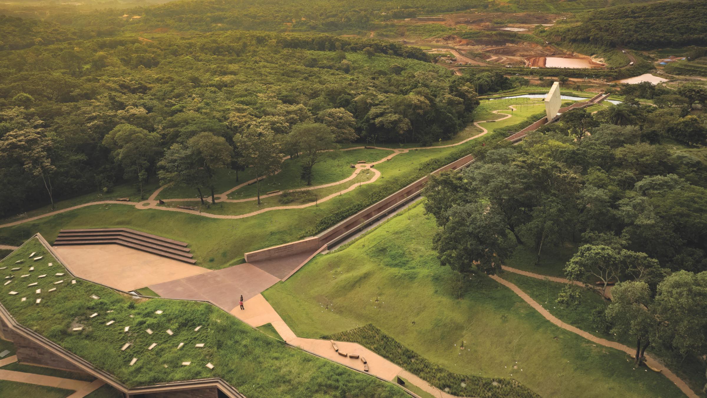 Aerial view of a modern building with geometric lines, surrounded by winding paths and dense green trees in a park-like landscape.