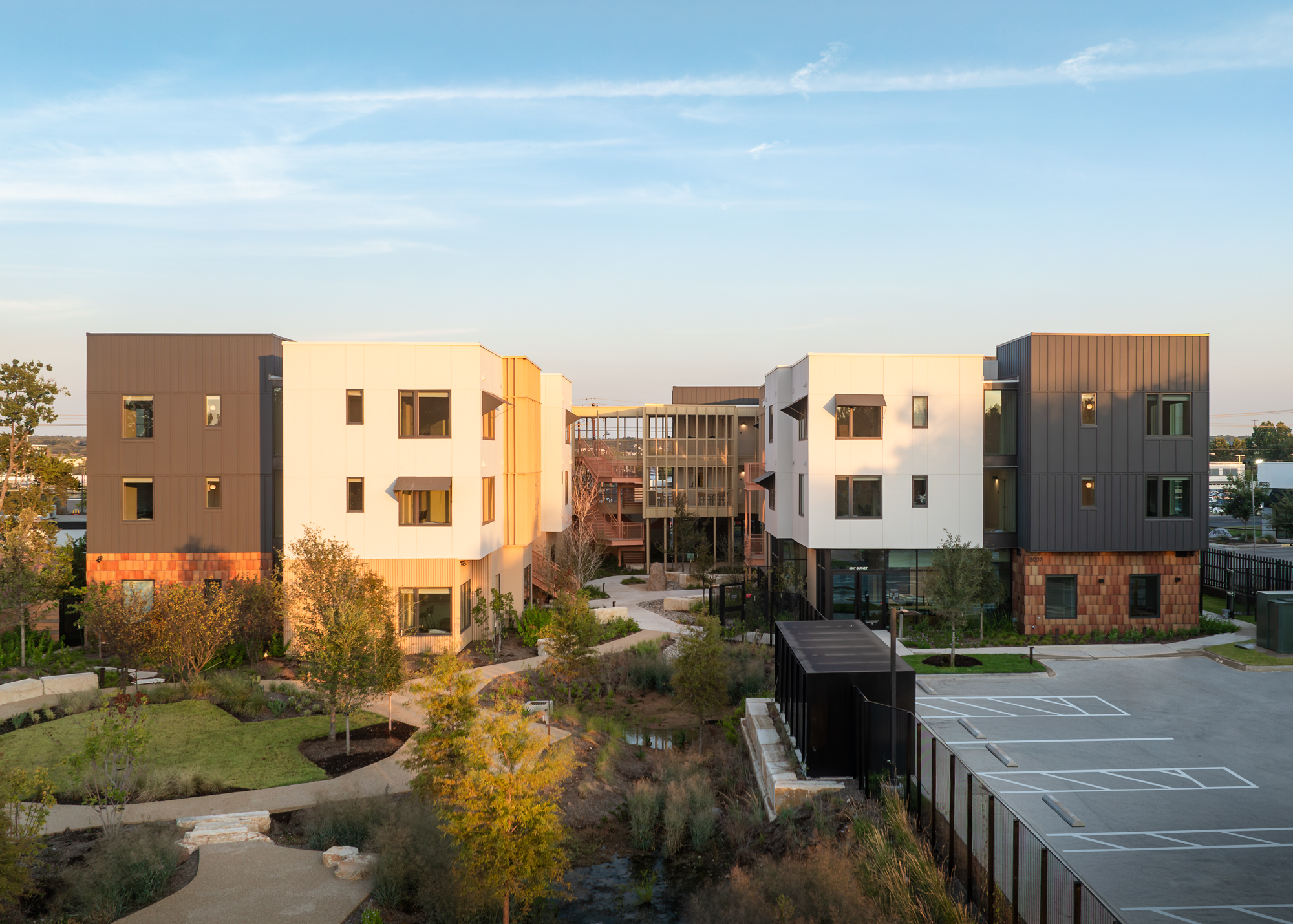 Modern three-story apartment complex with landscaped gardens, pathways, and an adjacent parking lot, photographed under clear skies.