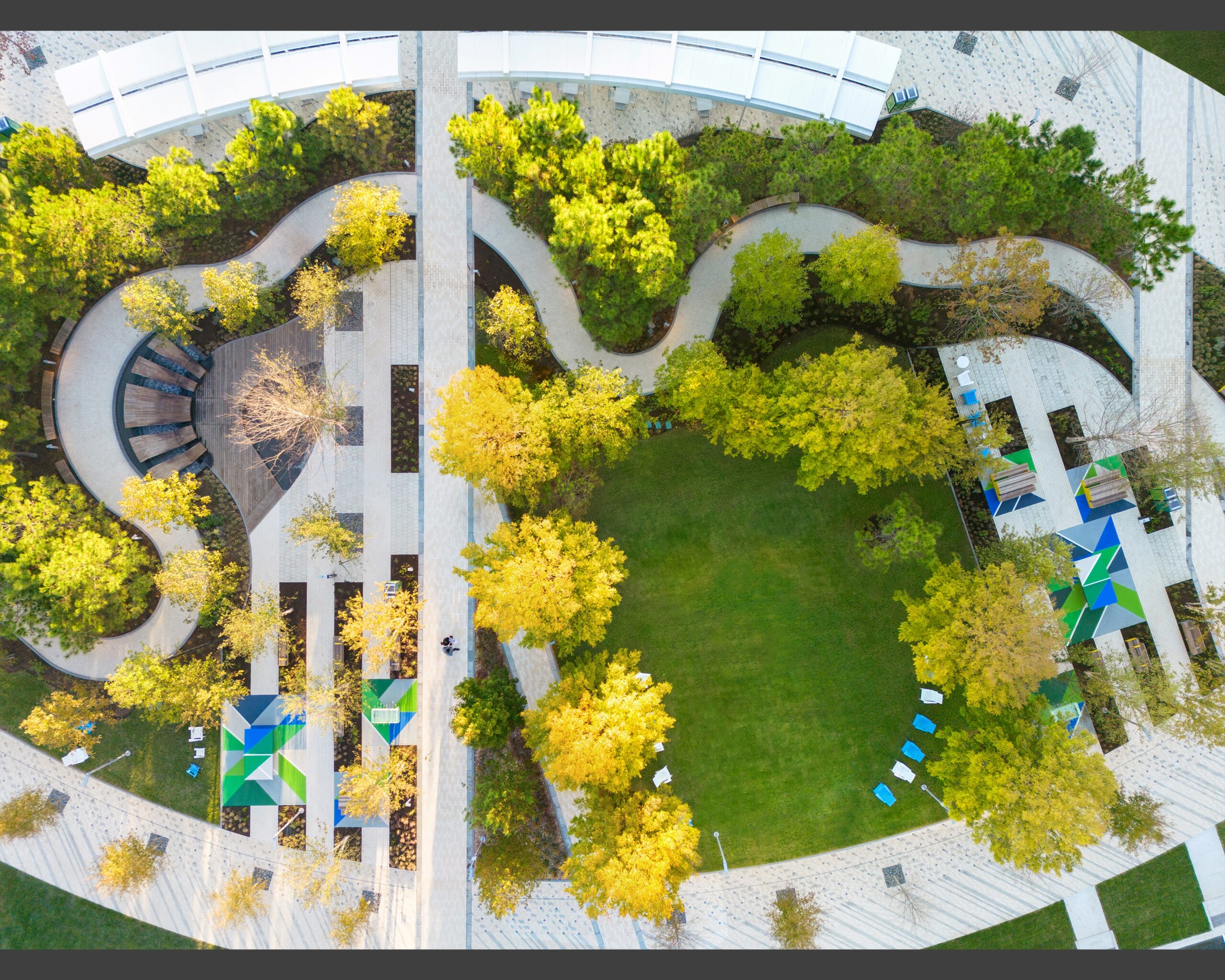 Aerial view of a landscaped park with winding paths, green lawn, trees, shaded seating areas, and geometric awnings.