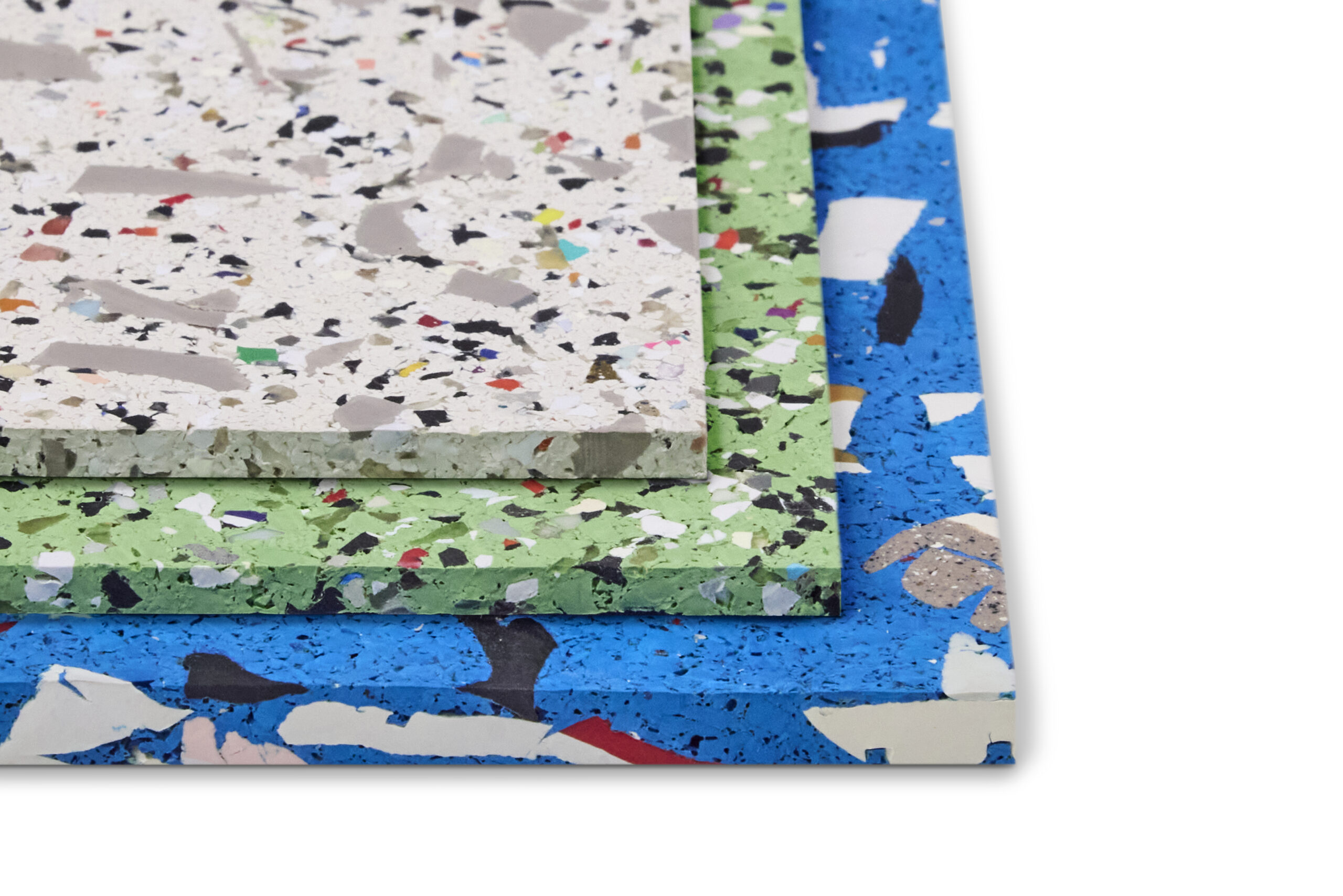 Three rectangular sheets of recycled composite material in white, green, and blue, each with a speckled, multicolored pattern, stacked in a corner on a white background.