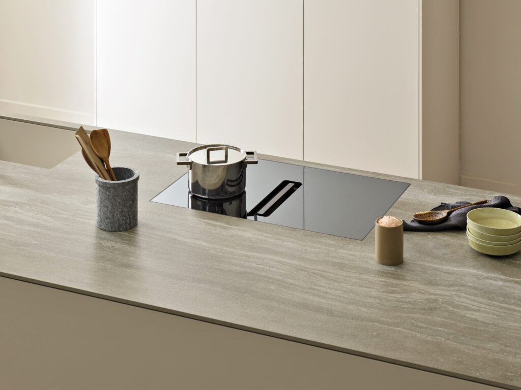 A silver pot sits on an induction cooktop on a kitchen island next to bowls, salt, and spoons. The island is a brown, marble-like surface and a beige cabinet sits in the background.