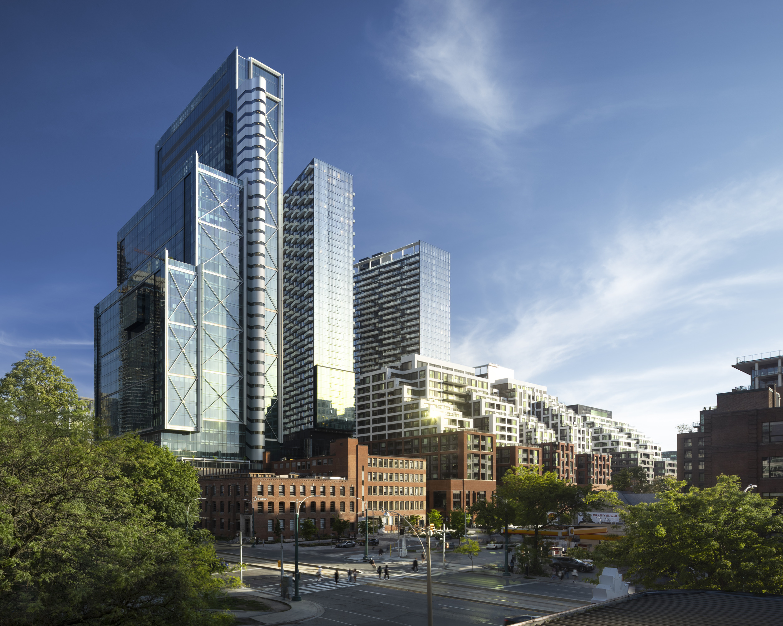 Modern glass skyscrapers stand behind older brick buildings on a sunny day, with a few trees and cars in the foreground and a clear blue sky overhead.