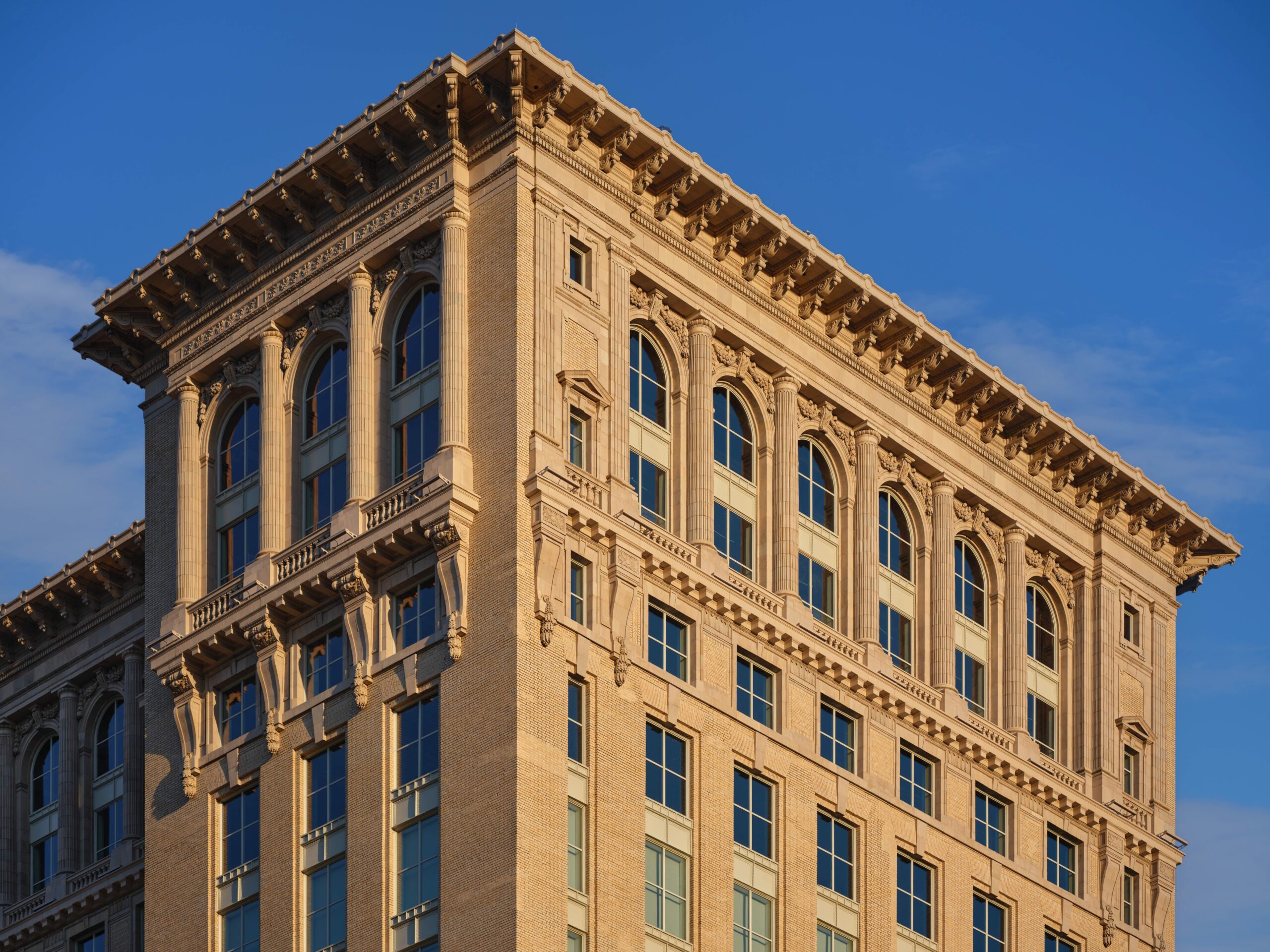 A beige, multi-story historic building with large arched windows and ornate architectural details, photographed against a clear blue sky.