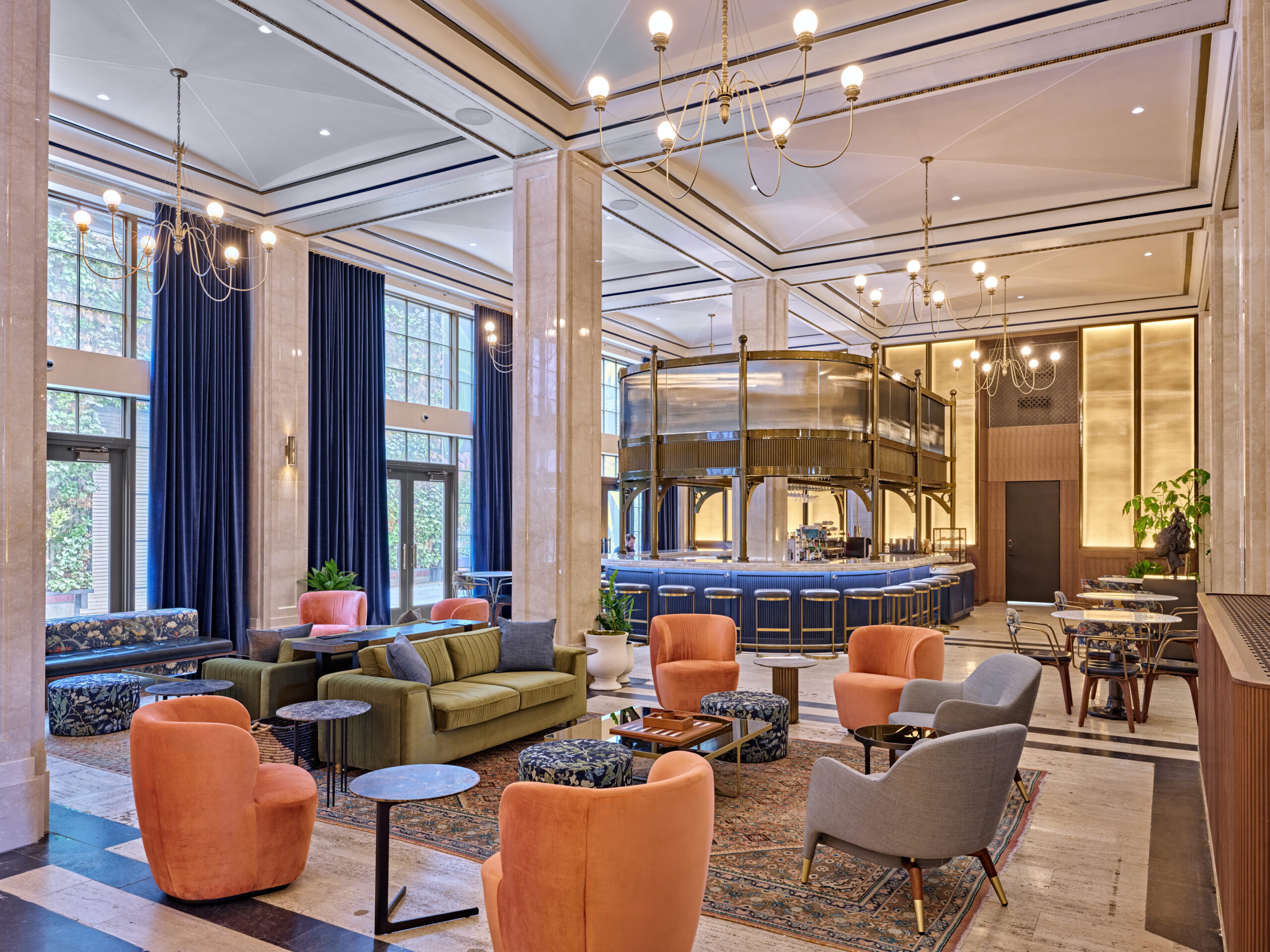 Spacious hotel lobby with modern seating, marble pillars, a central bar area, large windows, and chandeliers.