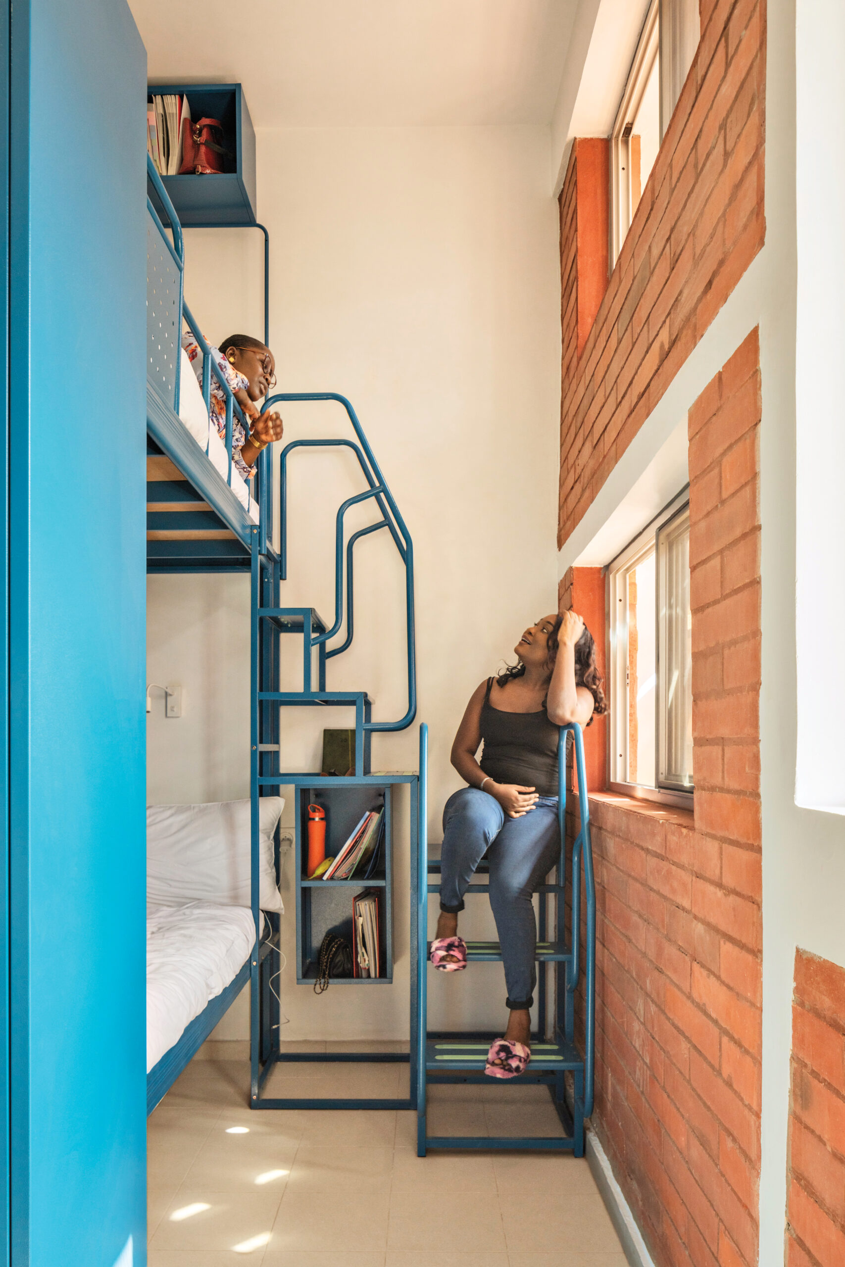 Two figures sit in a student bedroom that features a bright blue bunk bed with a modern staircase. One figure sits below, leaning agains the orange-brick wall, and the other looks down from the top bunk.