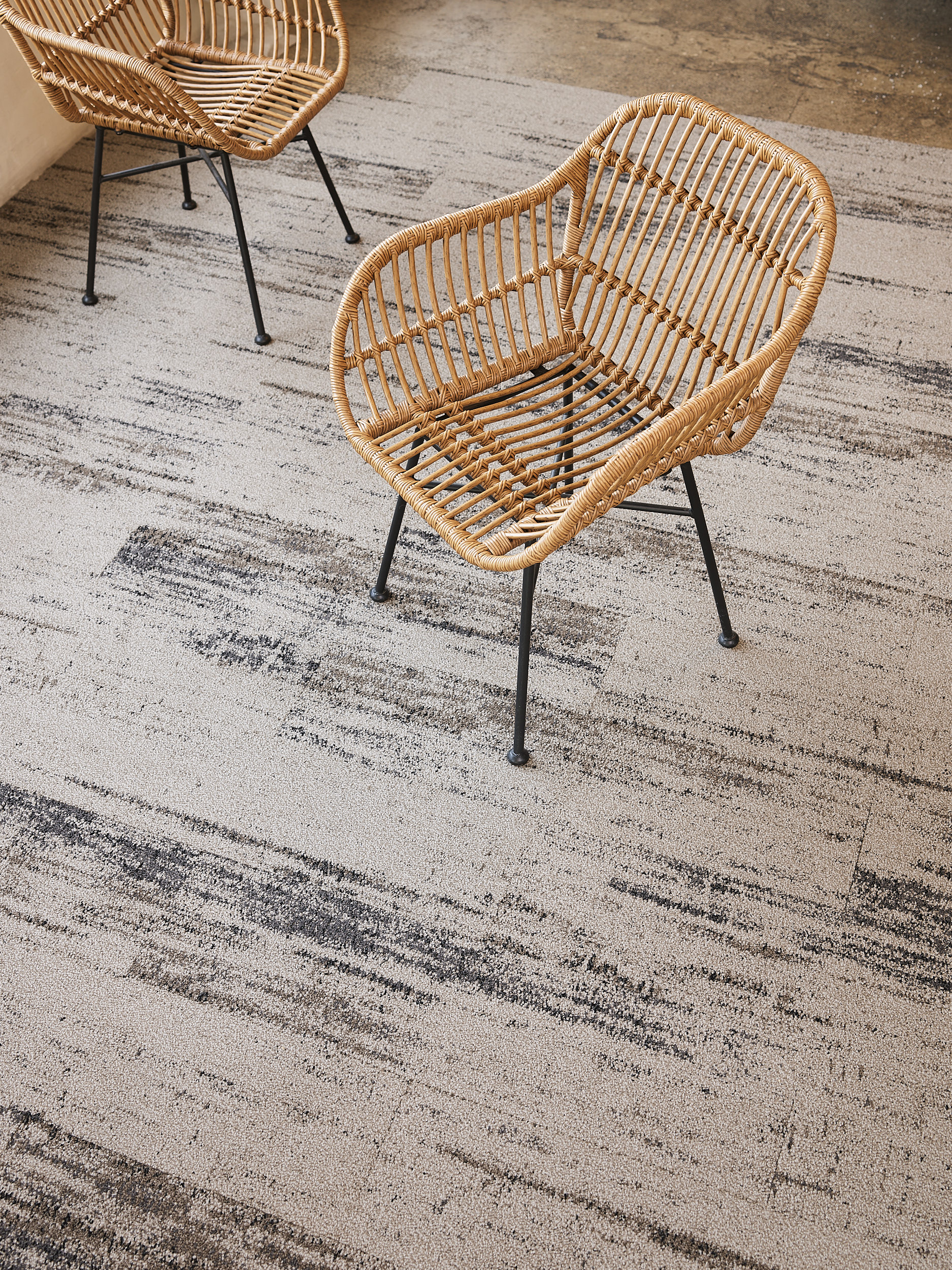 Two wicker chairs with black metal legs sit on a light gray, patterned carpet in a room with a concrete floor.