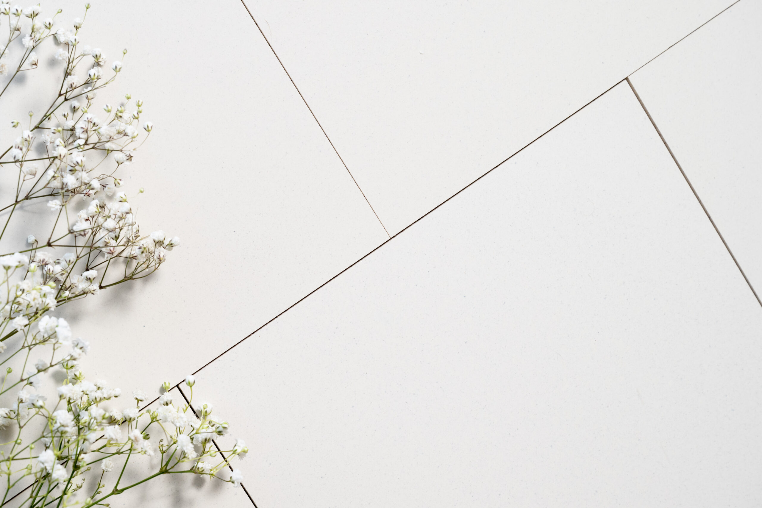 White tiles with thin grout lines are arranged diagonally. Small white baby’s breath flowers are placed in the lower left corner.
