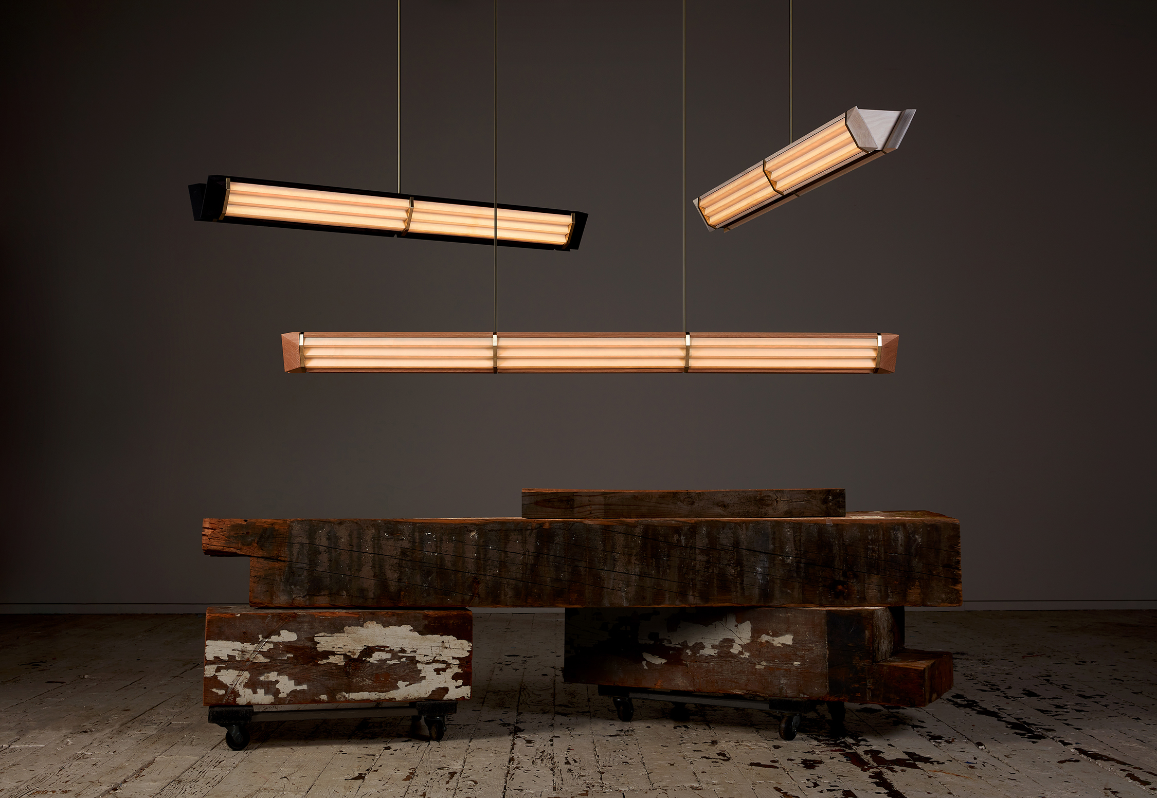 Three modern rectangular pendant lights hang above stacked, weathered wooden beams on casters, against a dark background and distressed wooden floor.