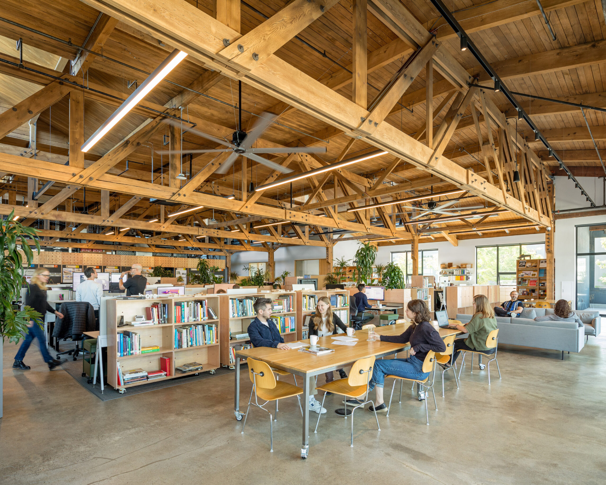 Open office space with exposed wooden beams, desks, bookshelves, and people working or meeting at tables and on a couch. Large windows provide natural light.
