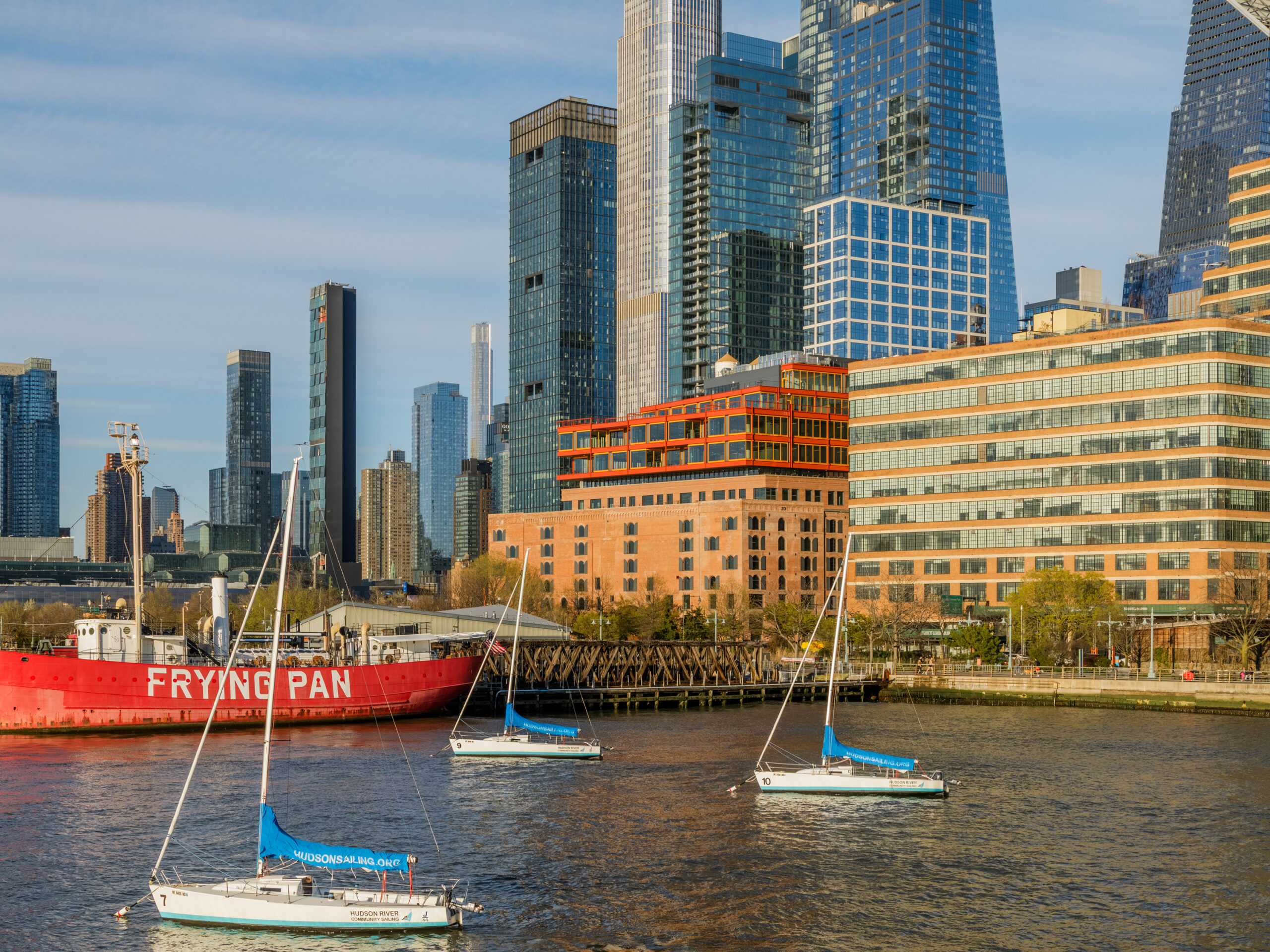 Three sailboats are on the water near a large red ship labeled “Frying Pan,” with modern high-rise buildings and offices in the background on a clear day.