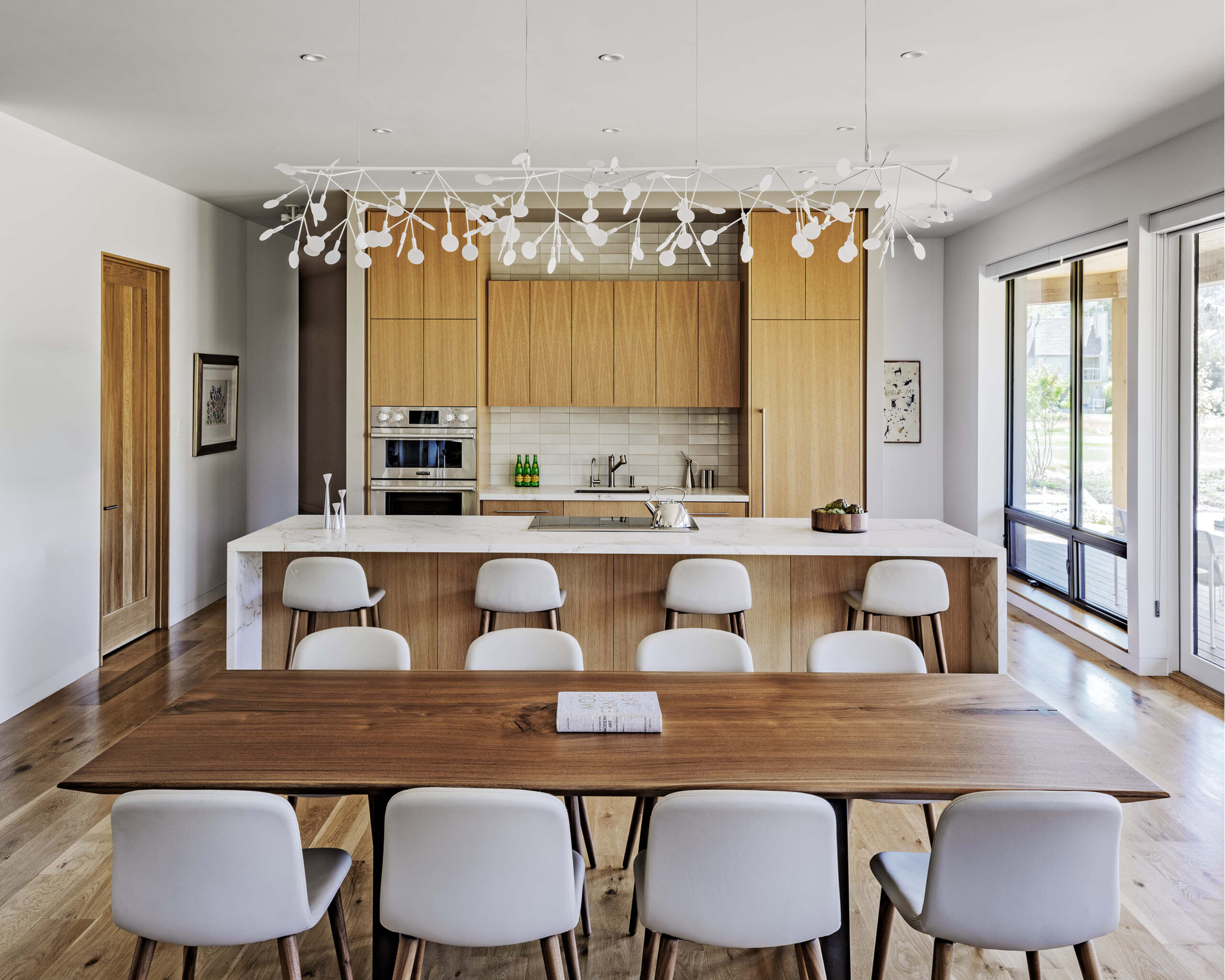 Modern kitchen and dining area with light wood cabinetry, a large island, a wooden dining table with white chairs, and a decorative white chandelier overhead.