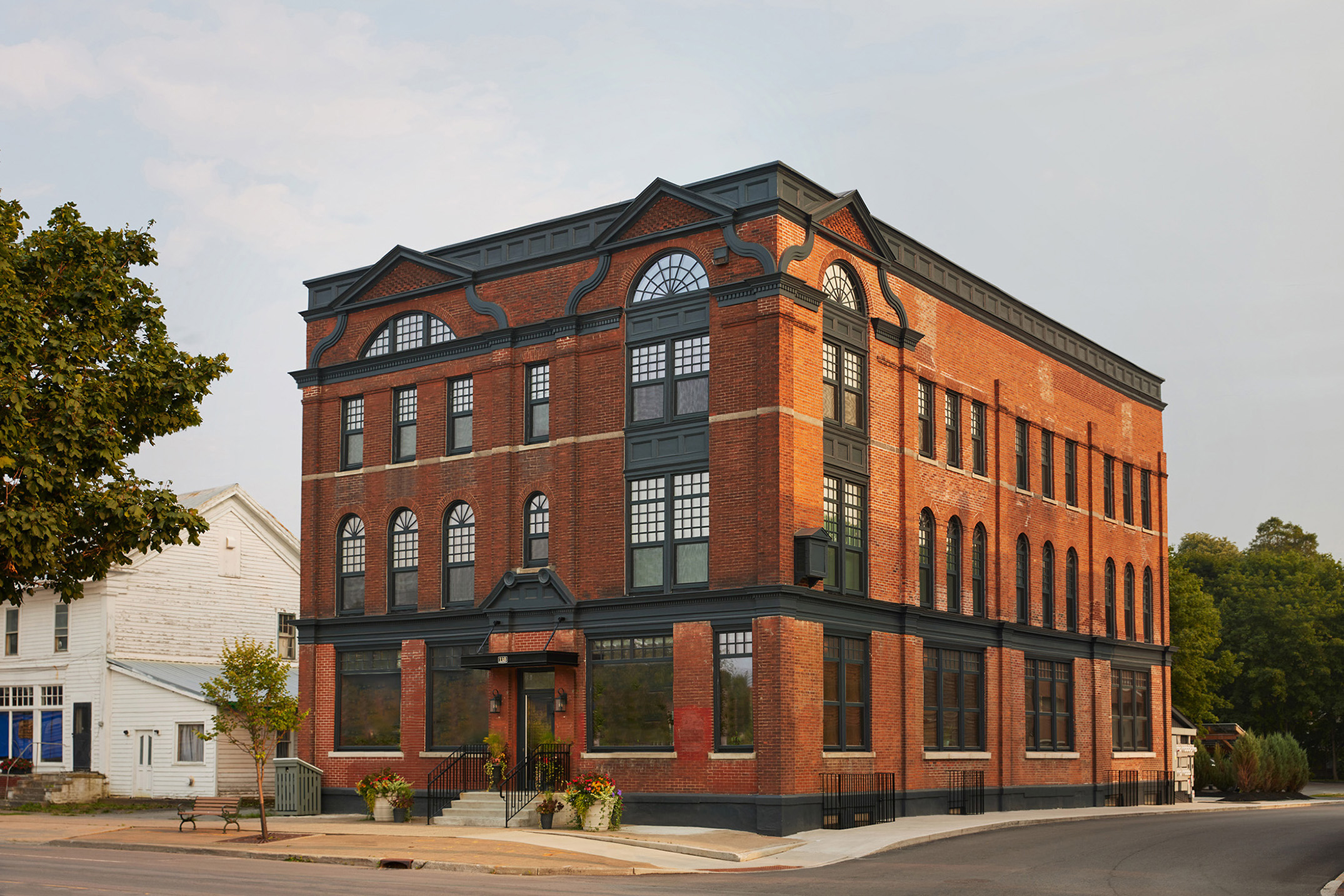 Three-story red brick building with large arched windows, black trim, and corner entrance, situated on a quiet street with trees and neighboring houses visible.