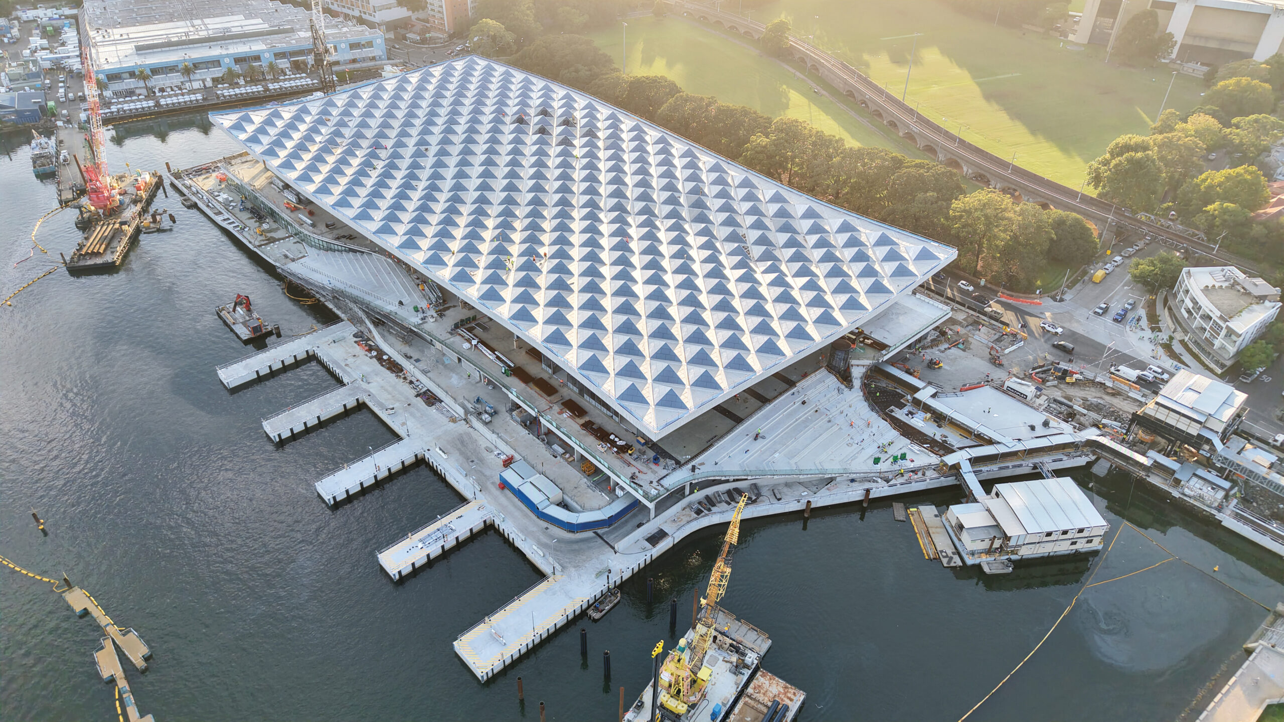 Aerial view of a large waterfront building by 3XN under construction with a distinctive geometric roof, surrounded by water, docks, machinery, and nearby roads.