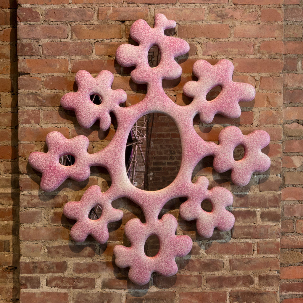 An irregularly shaped pink and white mirror with organic, blob-like edges is mounted on a red brick wall.
