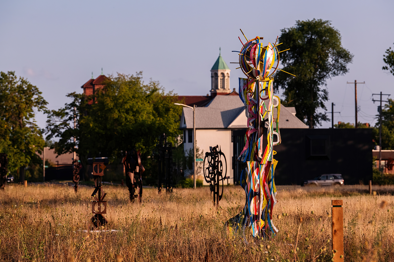 Several abstract metal sculptures stand in a grassy field, with a white church and trees in the background under a clear sky.