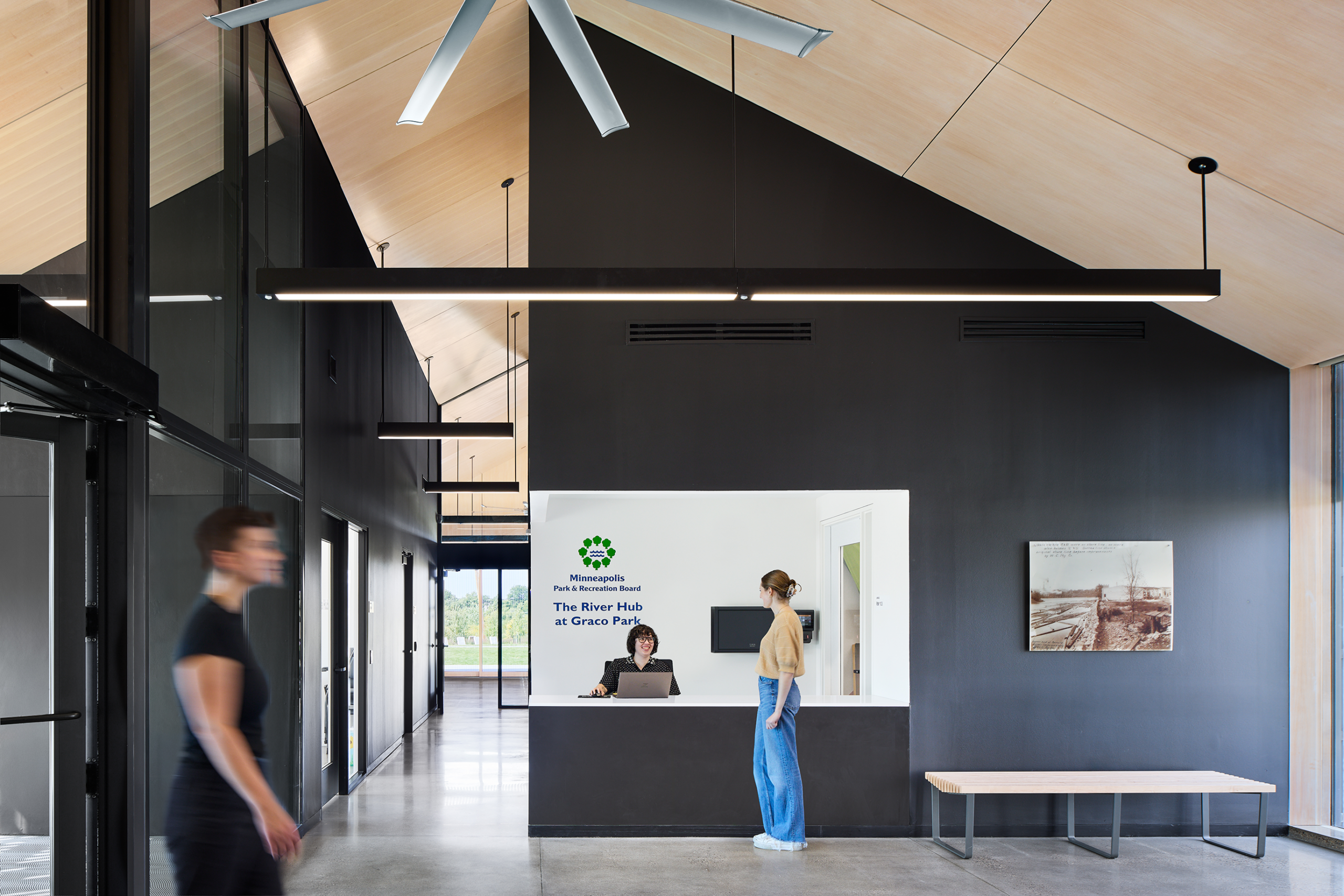 Modern reception area with high ceilings; a receptionist sits at a desk behind a white counter as a woman stands, and a person walks by in the foreground.