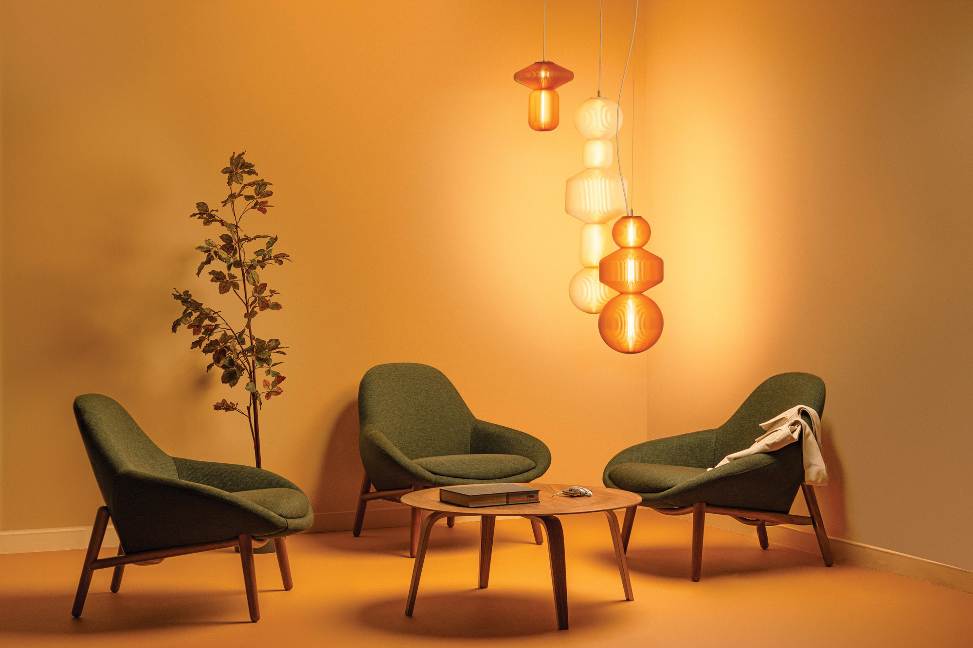 Three green armchairs surround a wooden coffee table with books and glasses, beneath modern pendant lights in a warm, orange-toned room with a small potted tree.