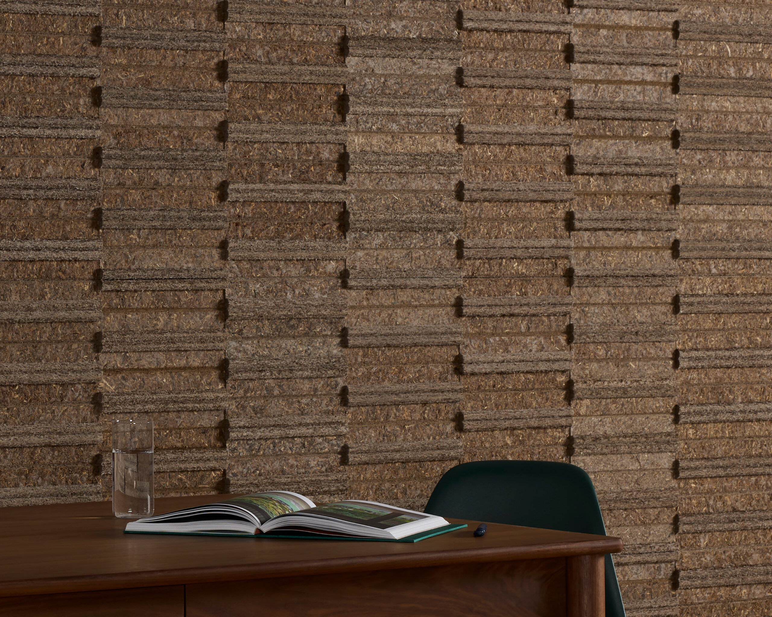 A wooden desk with an open book, a glass of water, and a dark chair, set against a textured brown wall with rectangular patterns.
