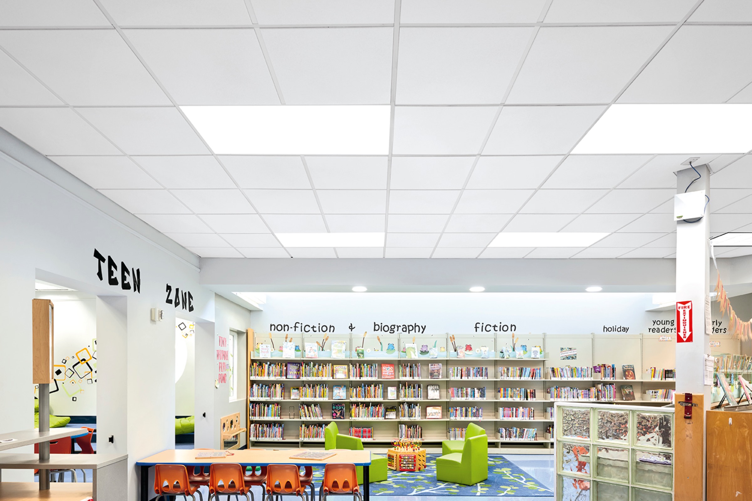 Colorful children’s library area with bookshelves, small tables and chairs, wall labels for book genres, and a green rug in the center under bright ceiling lights.