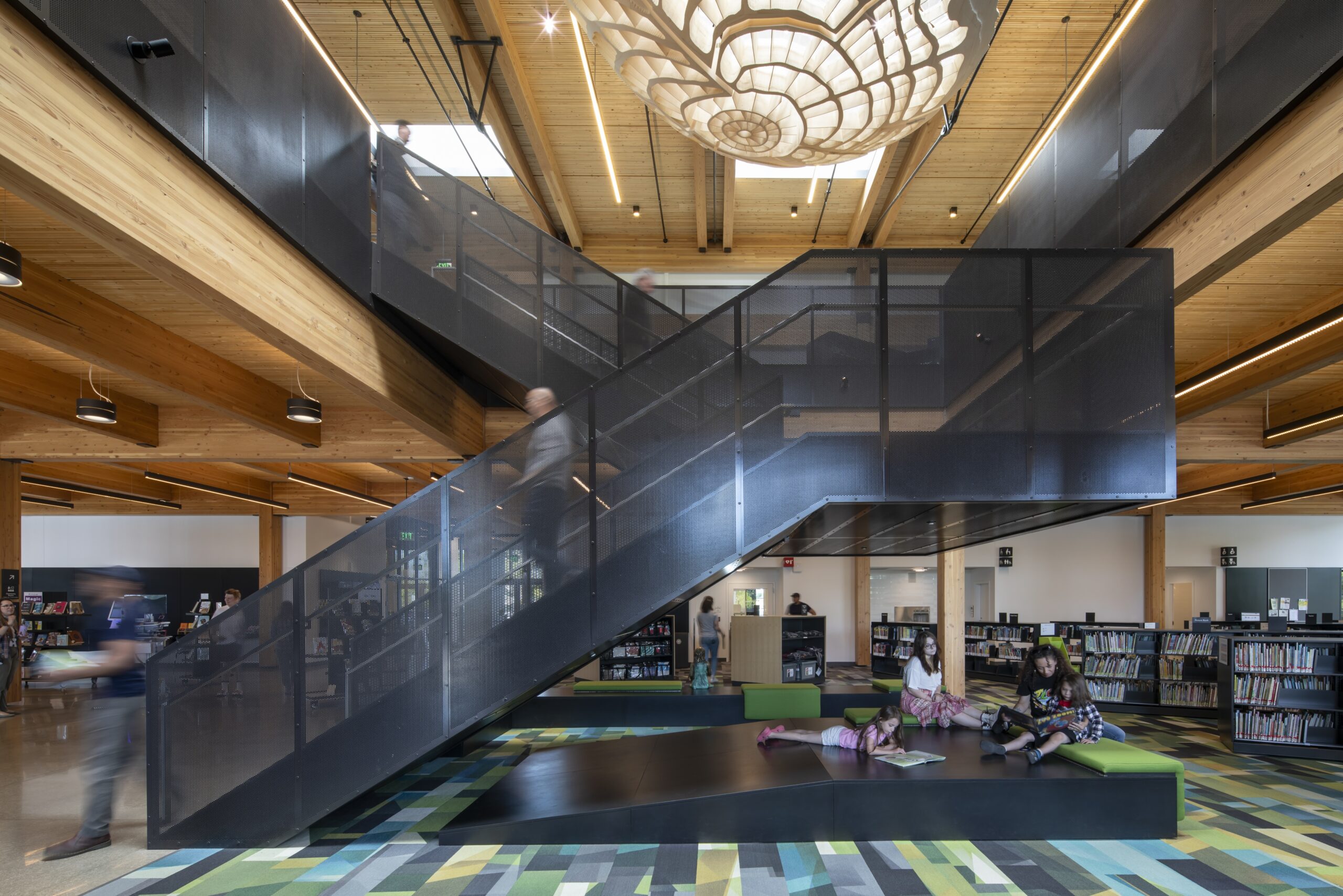 Modern library interior with a central black staircase, wooden ceiling, bookshelves, and children reading on green benches under geometric lighting.