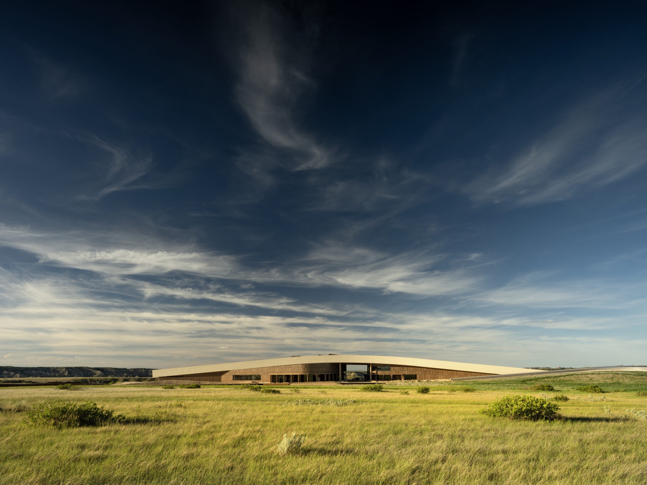 A modern, low-rise building sits in a wide grassy field under a dramatic sky with wispy clouds.