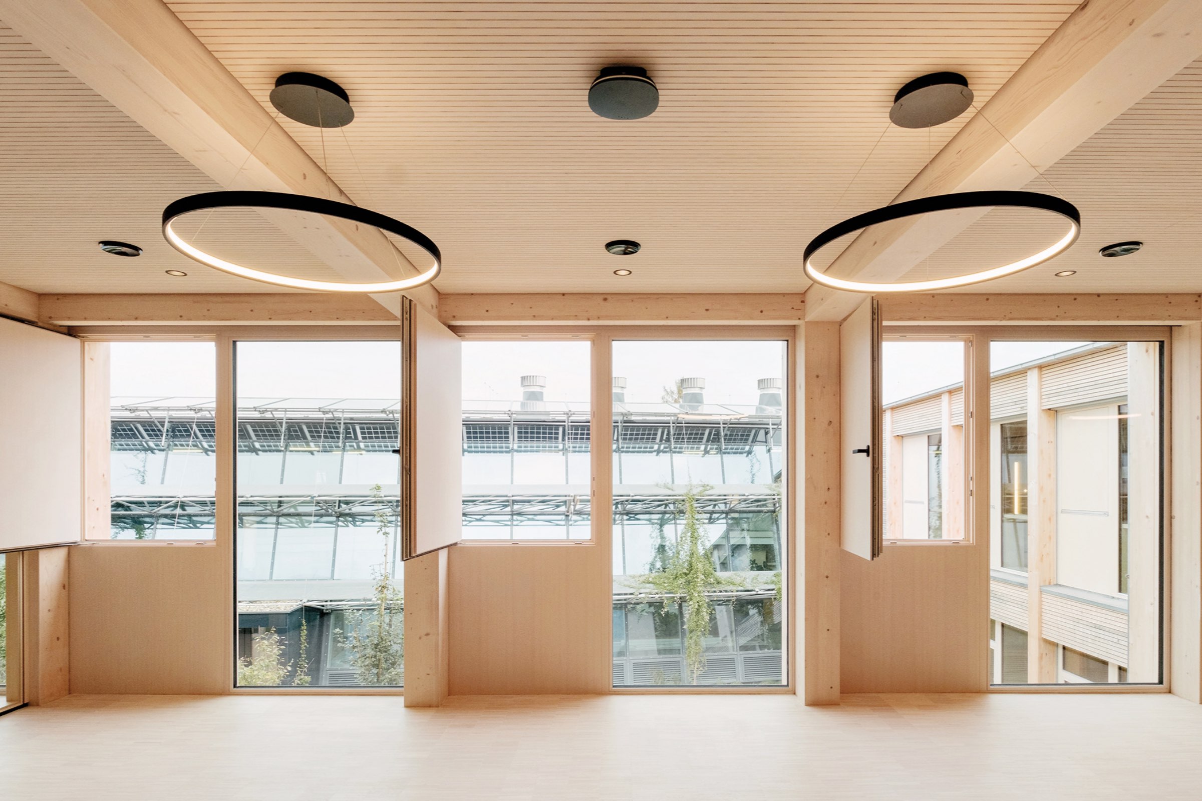 Modern interior by Juri Troy Architects with light wood ceiling and floor, large windows, circular LED lights, and open window panels facing an industrial building outside.