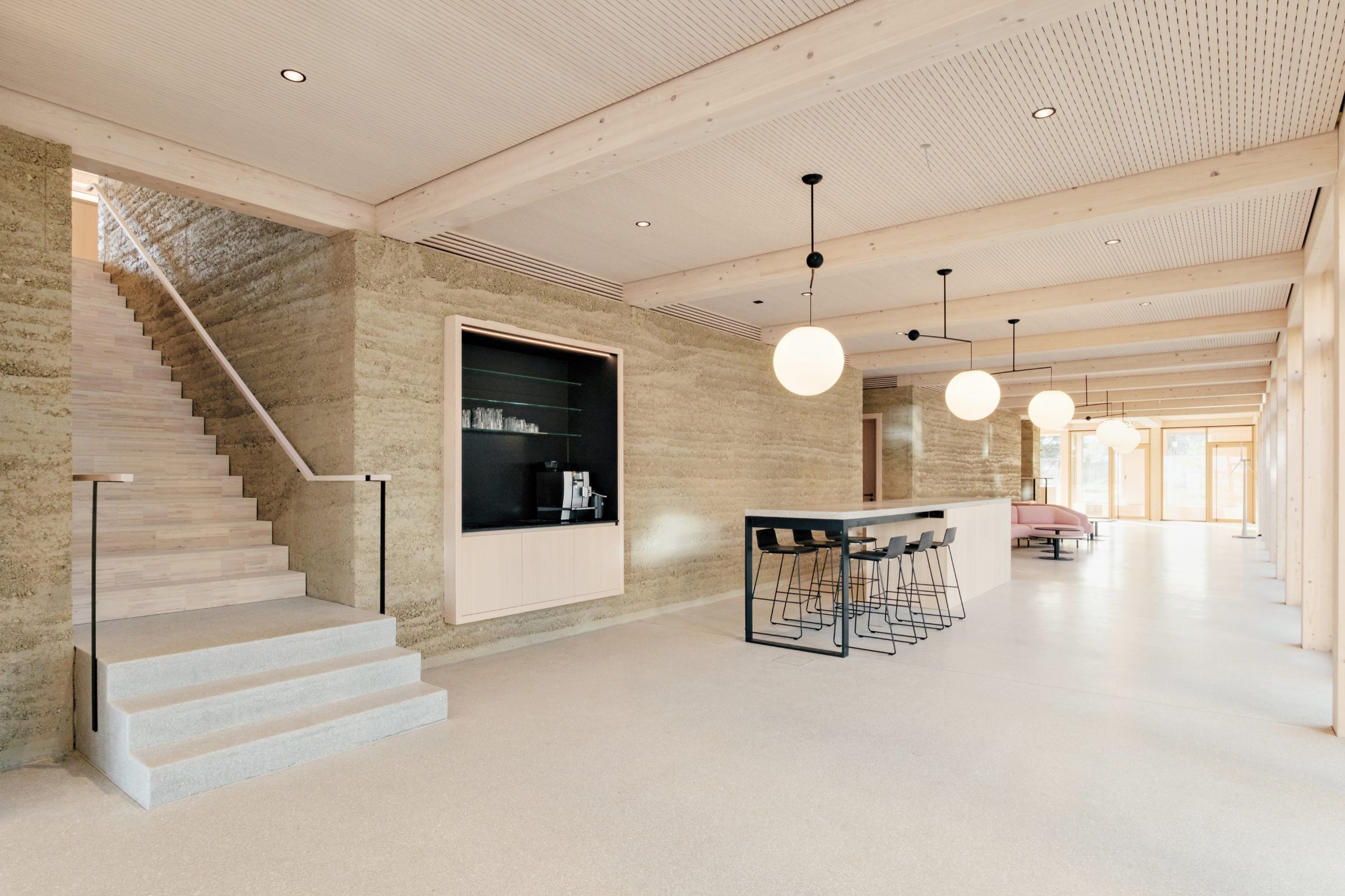 Modern open interior with light wood accents, a staircase on the left, a built-in coffee station, a bar with stools, and round ceiling lights. Large windows provide natural sunlight.