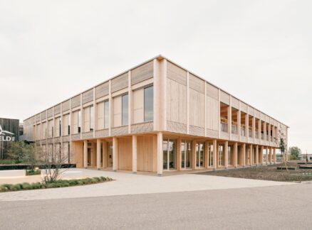 A modern two-story wooden office building with large windows, situated on a paved lot with minimal landscaping under a cloudy sky.