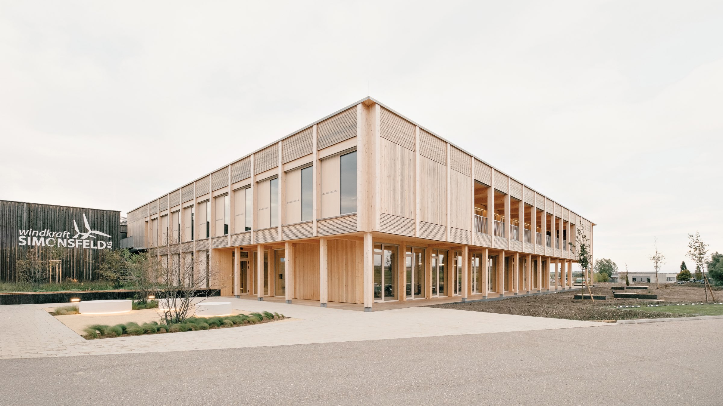 A modern two-story wooden office building with large windows, situated on a paved lot with minimal landscaping under a cloudy sky.
