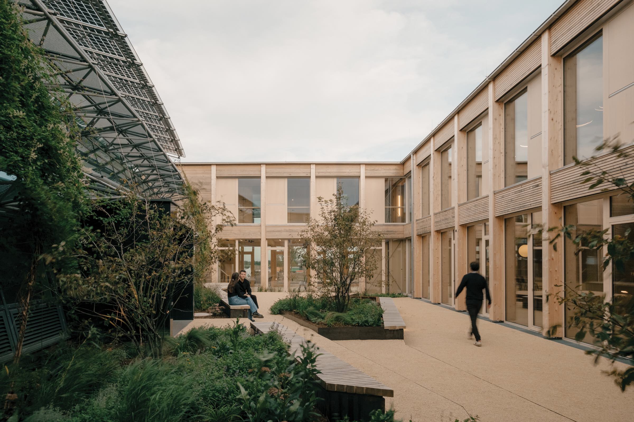 Modern courtyard by Juri Troy Architects surrounded by wood-paneled buildings, featuring greenery, two people—one seated, one walking—and large windows under a partly cloudy sky.