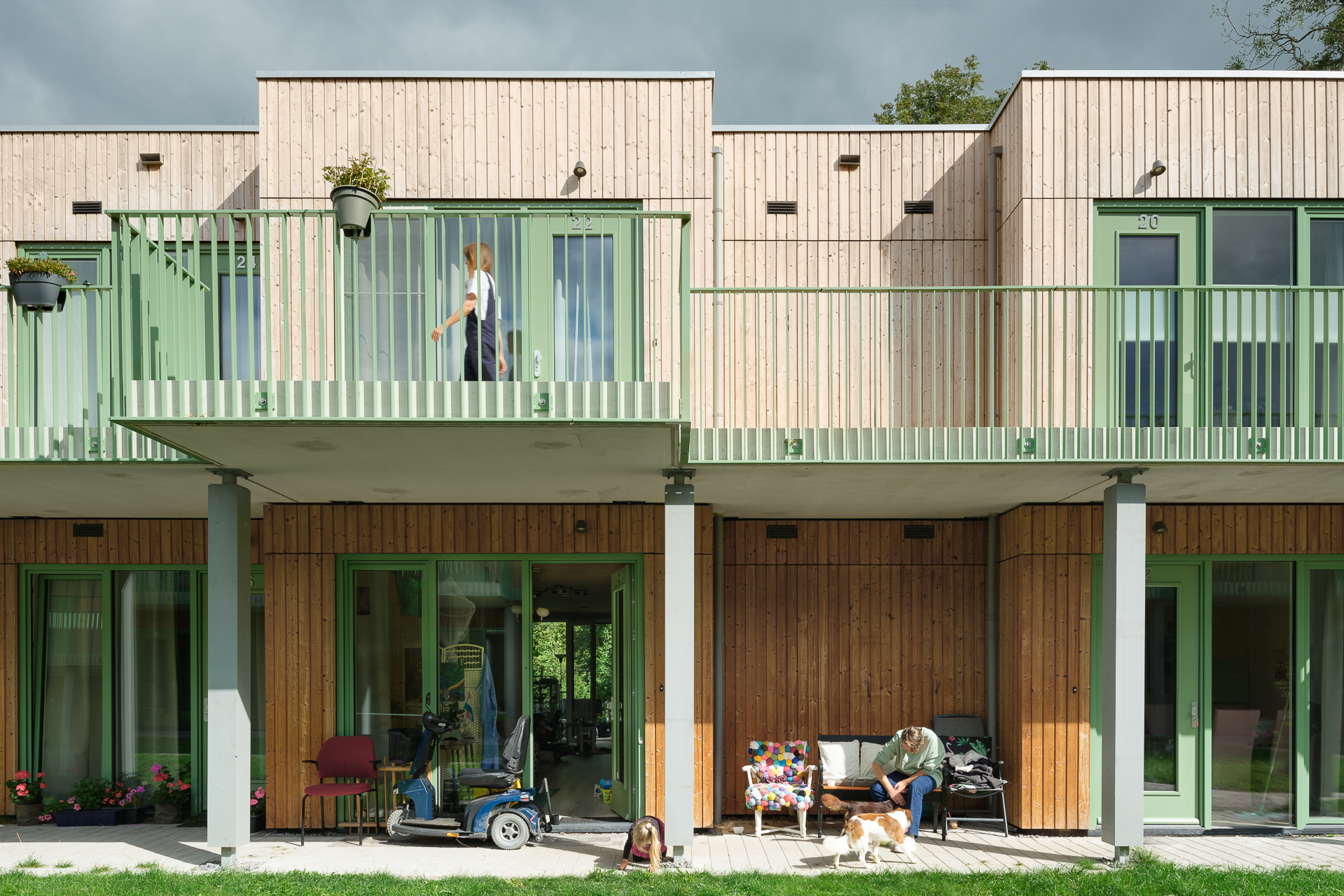 Two-story wooden apartment building with green trim; a person stands on the balcony above while another sits below with a dog. Potted plants and various items are on the patio and balcony.