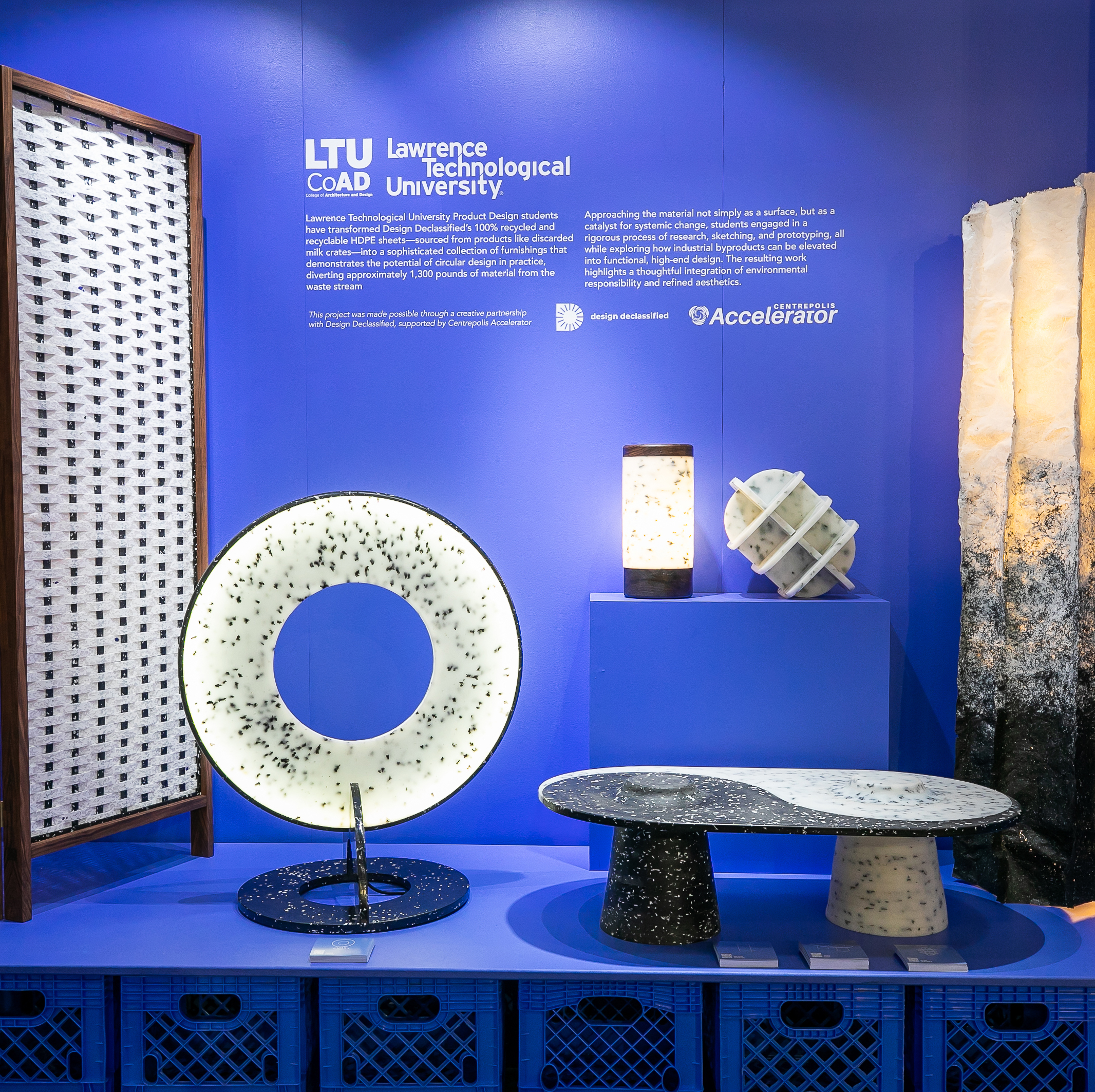A display of modern furniture and lamps with speckled designs is arranged against a bright blue wall bearing Lawrence Technological University’s logo and descriptive text.