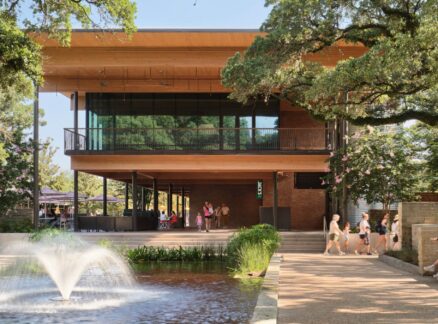A modern two-story building with glass walls, designed by Lake Flato, overlooks a pond with a fountain; people walk and sit on benches under large trees on a sunny day.