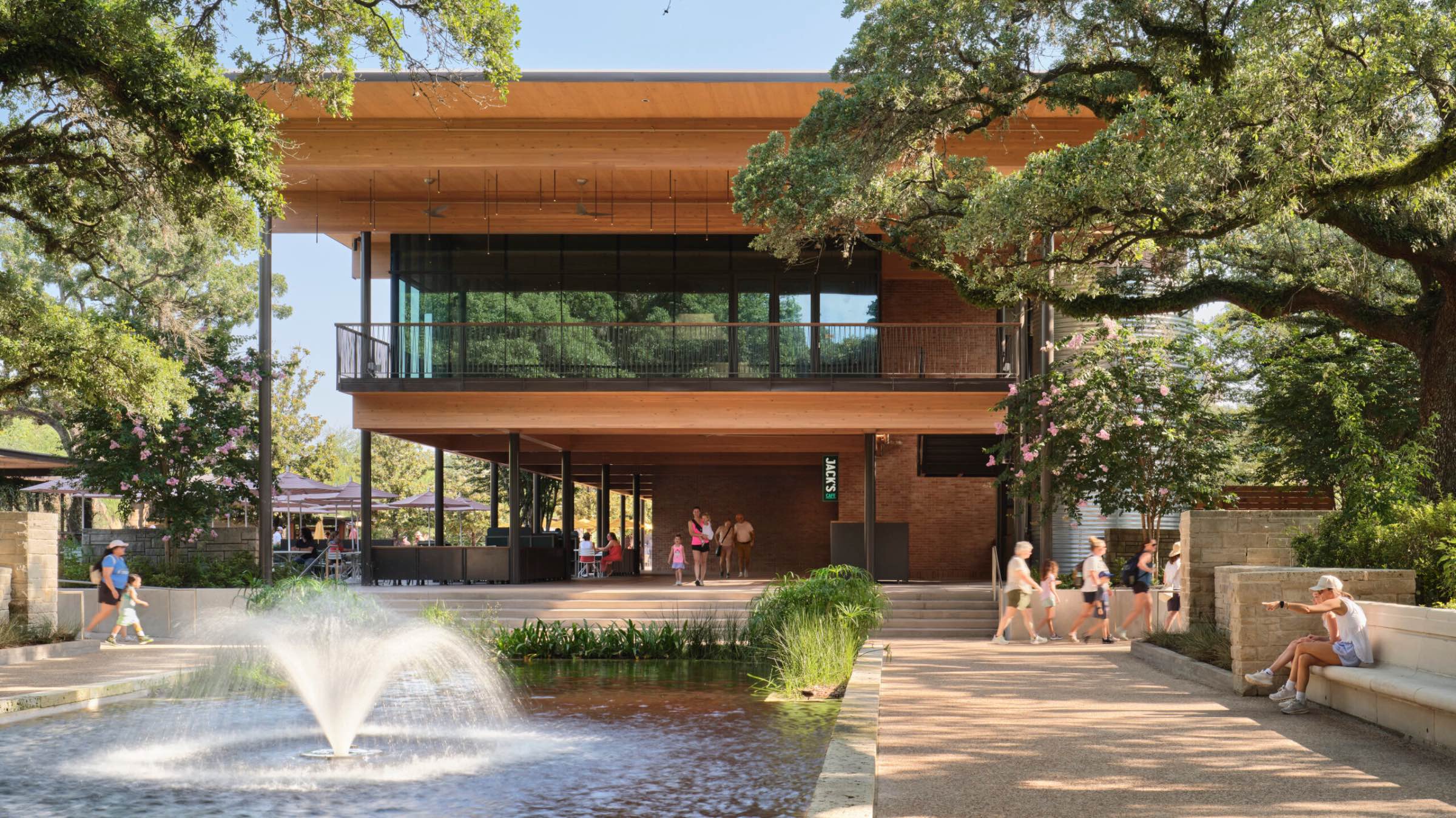 A modern two-story building with glass walls, designed by Lake Flato, overlooks a pond with a fountain; people walk and sit on benches under large trees on a sunny day.