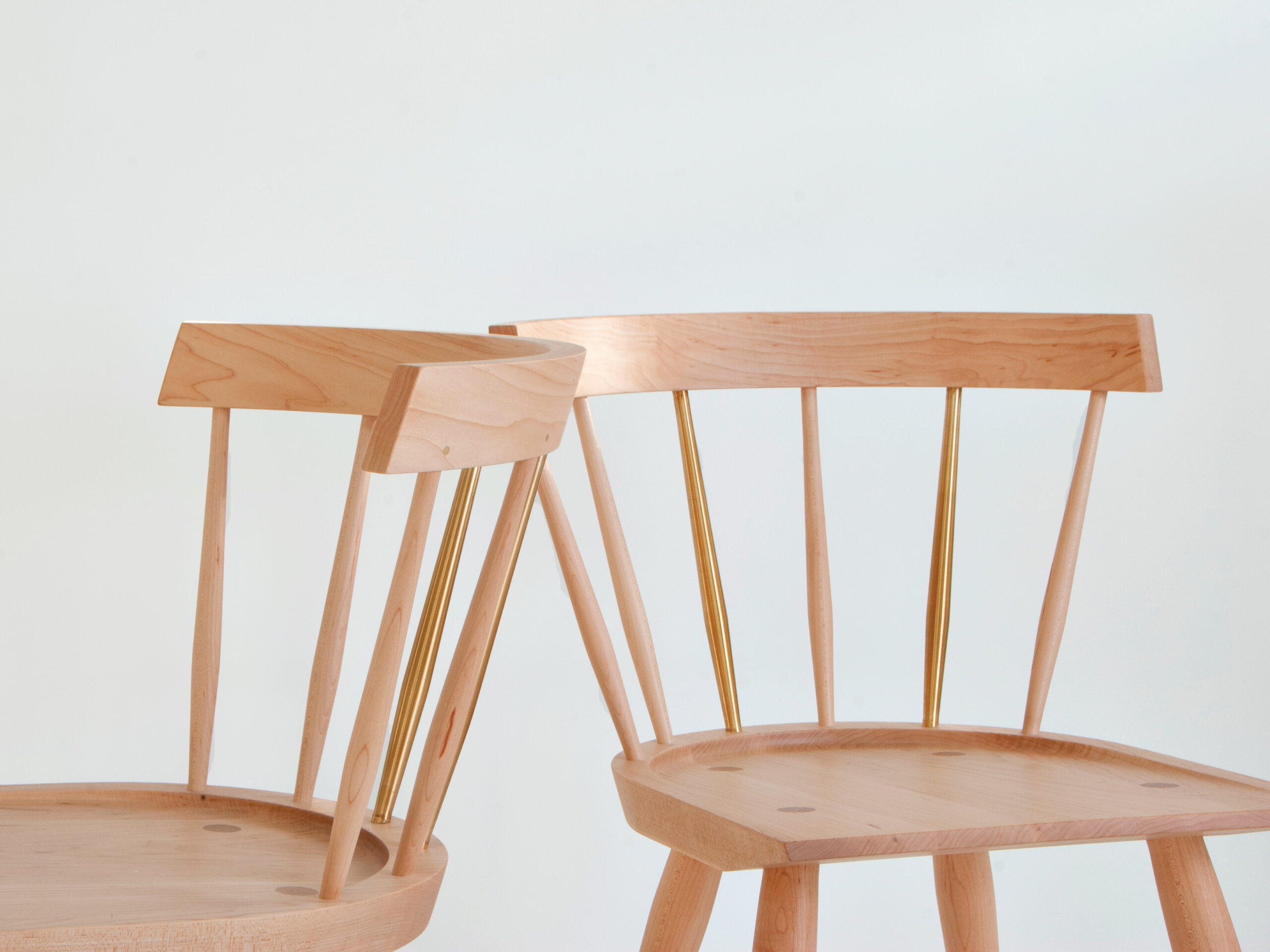 Two light wood chairs with spindle backs and gold-toned rods are placed against a plain white background. The chair on the left is partially leaning against the chair on the right.