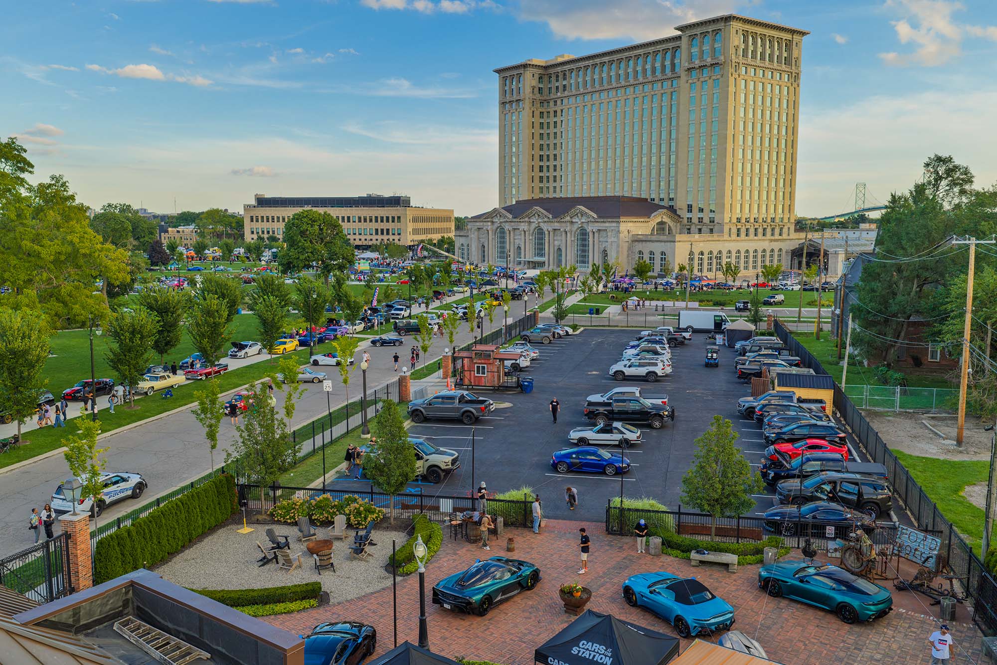 A parking lot filled with cars near a large historic building, surrounded by trees and people walking around, under a blue sky.