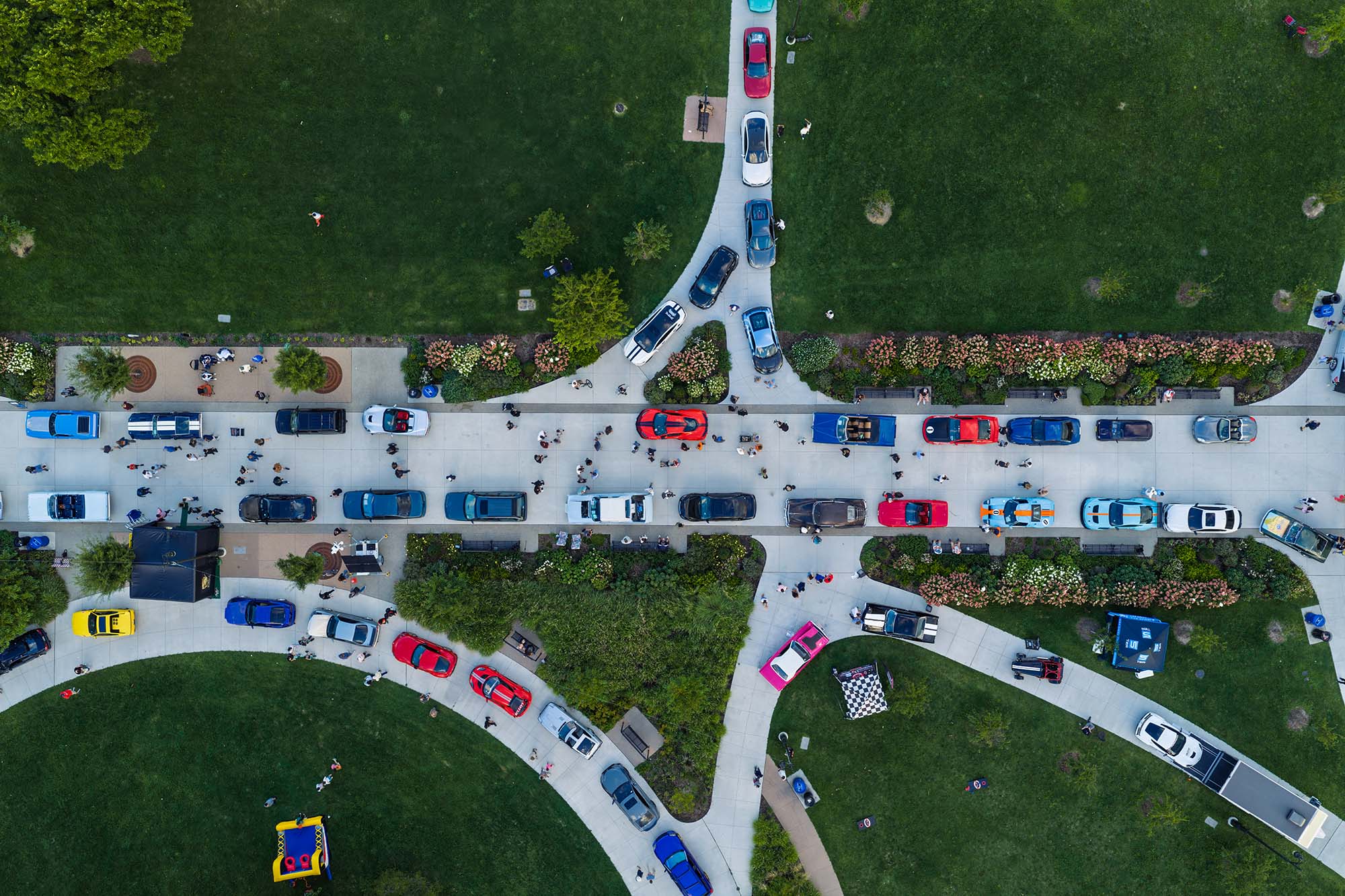 Aerial view of cars parked along a street with people walking on sidewalks in a park-like setting with green lawns and pathways.