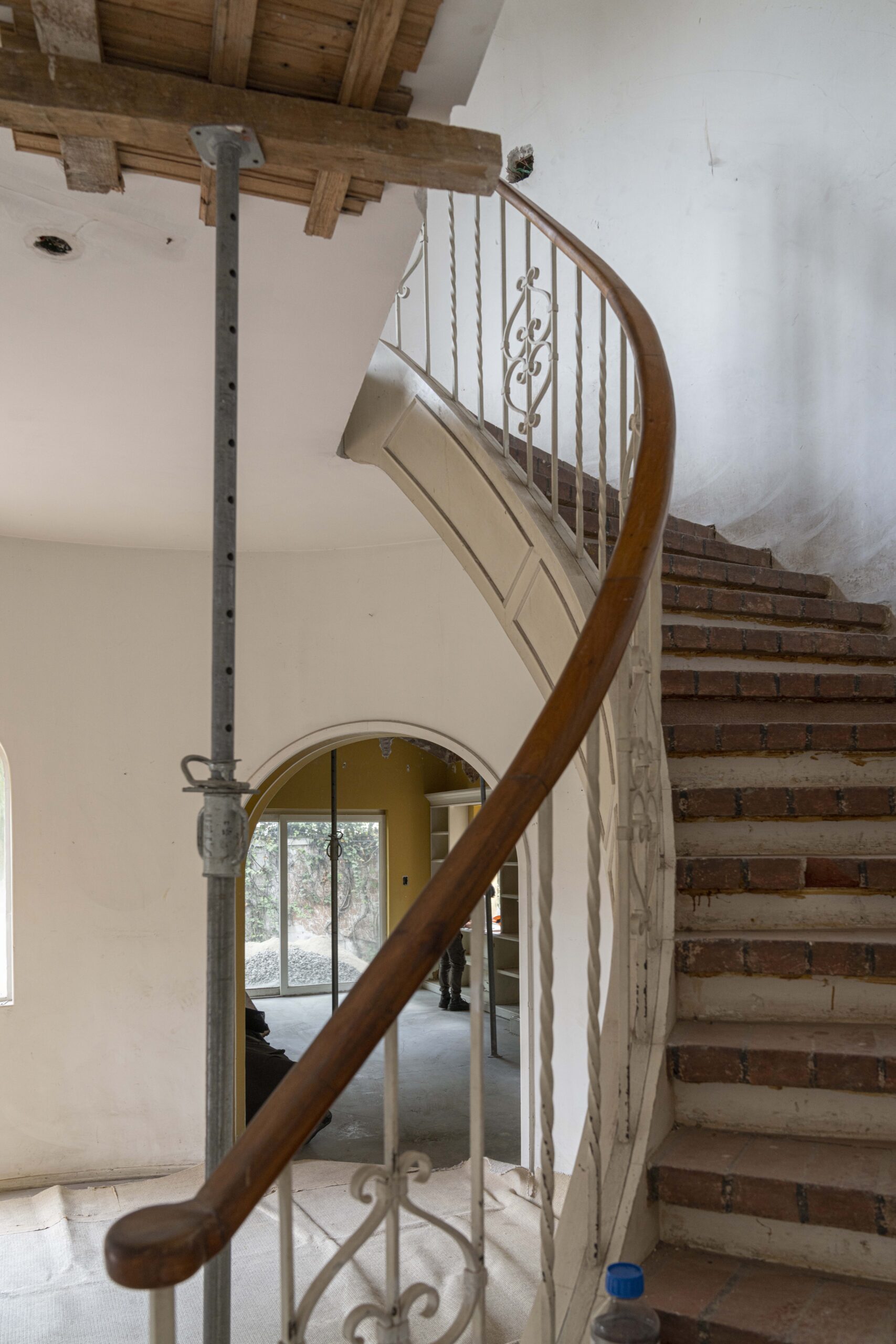 A curved staircase with brick treads and a wooden handrail under renovation, with a metal support pole in the foreground.